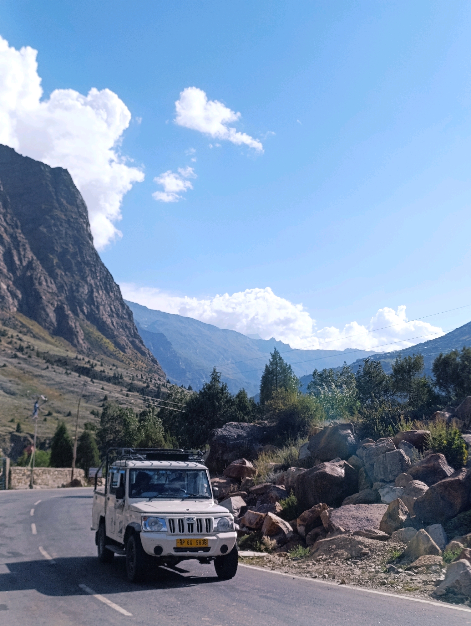 In Gemur, India, a white Mahindra Bolero pickup truck is parked on the right side of a paved road that curves to the left. The truck, facing slightly away from the viewer, has a roof rack and a yellow license plate that reads "HP 66 5838". The road is lined with large rocks and vegetation on the right, and on the left, a steep, barren mountain slope rises dramatically. In the background, successive mountain ranges extend into the distance under a bright blue sky with scattered white clouds. The scene captures a remote, mountainous landscape under daylight conditions.
