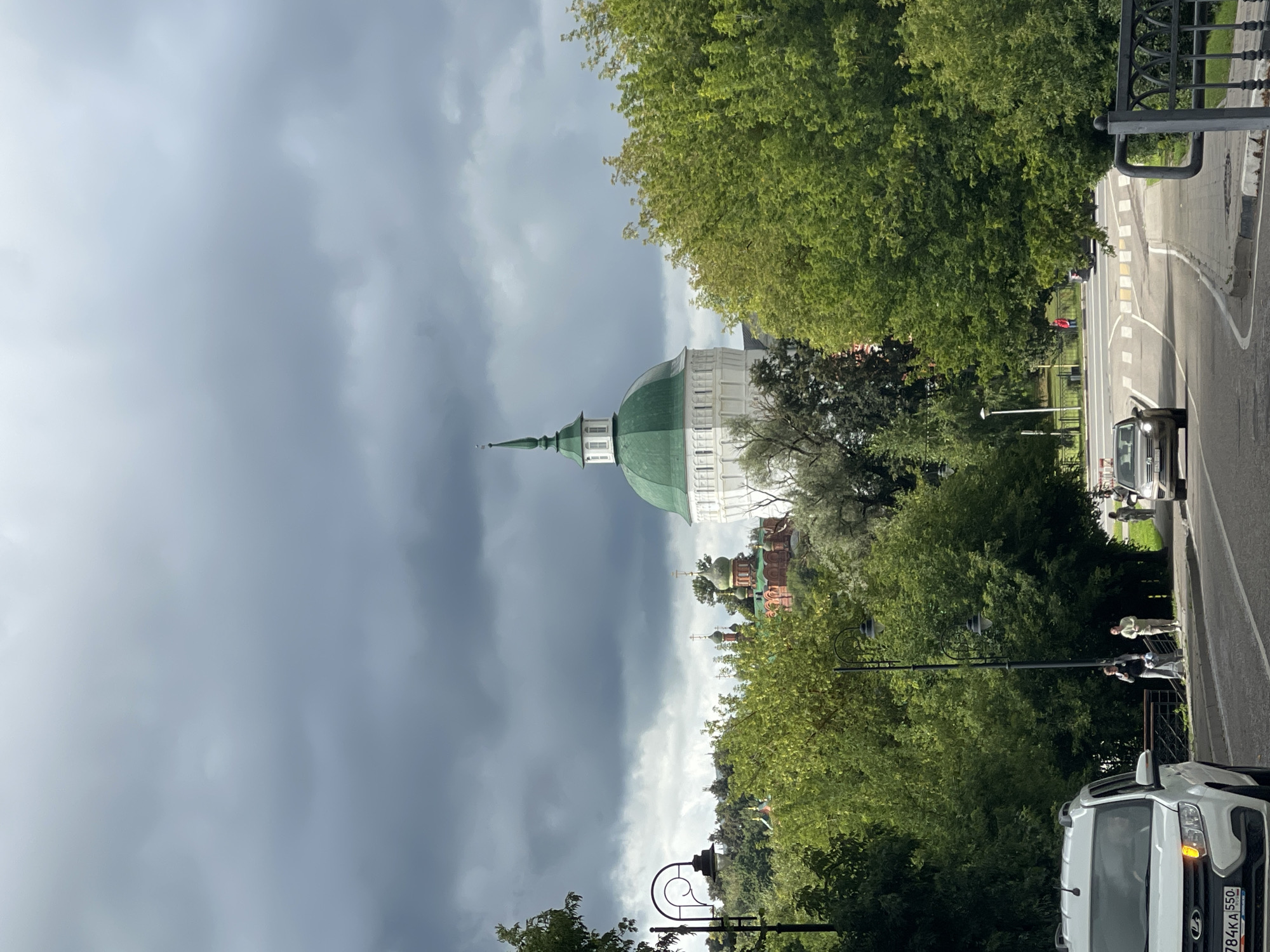The image shows a view of the Trinity Lavra of St. Sergius in Sergiev Posad, Russia, under a cloudy sky. In the foreground, a white car is parked on the left side of the road, and a silver car is driving away from the camera on the right. Several people are walking on the sidewalk to the right of the road. Lush green trees line the street and surround the buildings of the monastery. The monastery itself features several distinct structures, including a prominent white building with a green dome and spire, and other buildings with red brick and smaller green domes, characteristic of Russian Orthodox architecture. The overall scene depicts a moment in a city with historical and religious significance, captured on an overcast day.