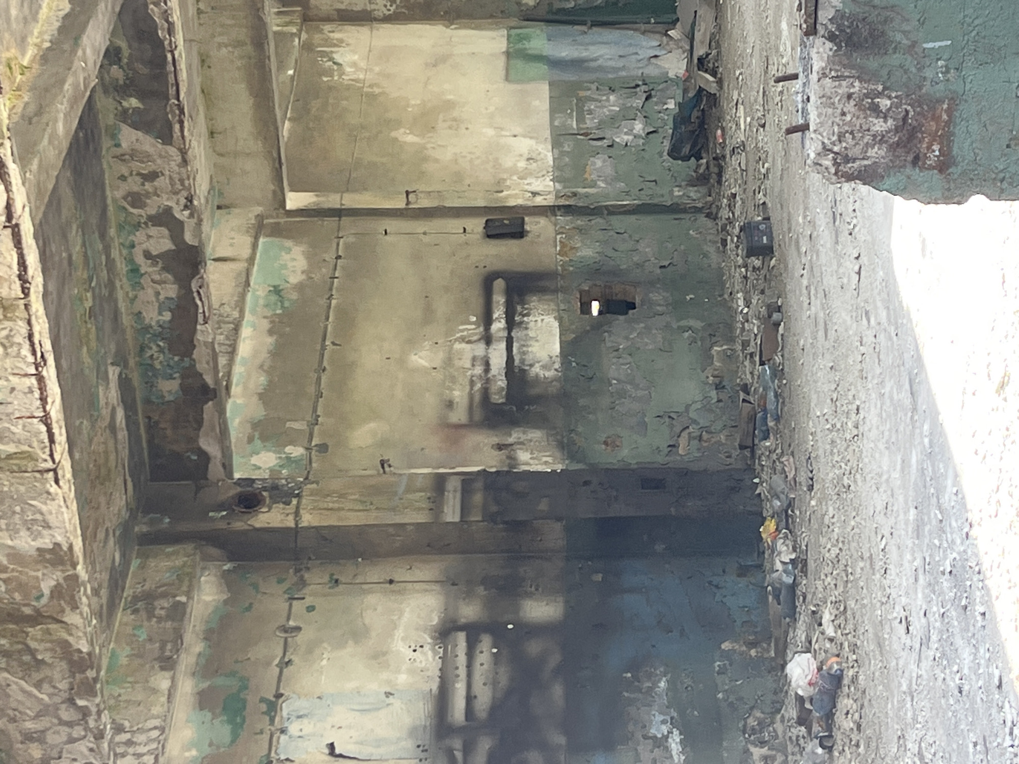 The image shows the interior of an abandoned building in Burgas, Bulgaria. The walls are concrete and covered with peeling paint in shades of gray, green, and white. There are rectangular holes and dark stains on the walls, possibly from fire damage. Debris is scattered along the floor, especially near the right side of the image where the wall meets the floor. The building appears to be in a state of disrepair, with exposed rebar and crumbling concrete. Natural light streams in from an opening on the right side of the image.