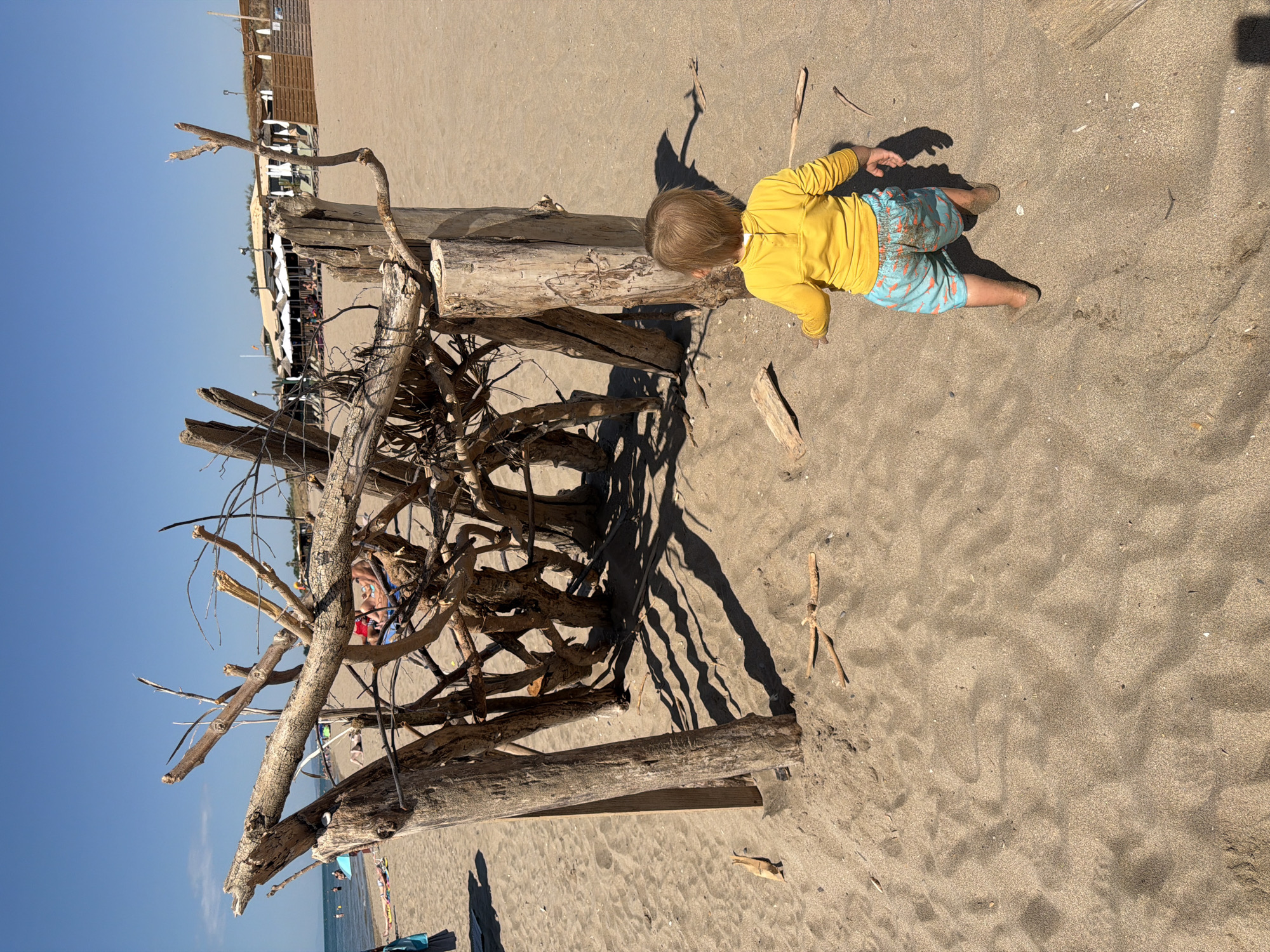 A toddler wearing a yellow long-sleeved shirt and patterned swim trunks stands on a sandy beach in Sérignan, France. The child is facing away from the camera, looking towards a structure made of driftwood and logs. In the background, the ocean is visible with a clear blue sky above. Further back, there are beach umbrellas, lounge chairs, and some buildings, suggesting a beach resort or establishment. The sand is textured with ripples and scattered pieces of driftwood.