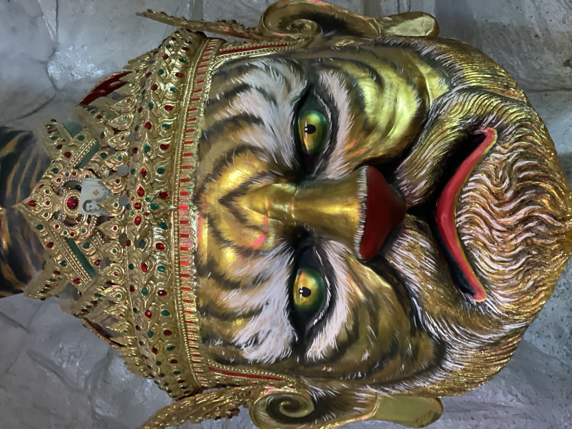 A close-up, head-on view of a golden, painted mask depicting a stylized tiger face, adorned with an elaborate crown. The mask features bold black stripes on a golden face, white fur details around the eyes and muzzle, bright green eyes with black pupils, and a red nose and mouth. The crown is ornate, with red and green jewels and intricate gold detailing. A small, framed portrait of a man is affixed to the front of the crown. The background is a textured, light-colored surface, possibly a wall. The image appears to be taken indoors. This artifact is located at the Wat Phra That Doi Suthep, a temple in Chiang Mai, Thailand.