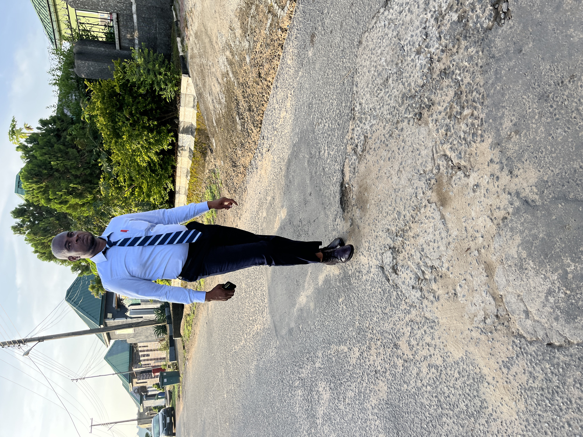 An adult male stands outdoors on an uneven, textured asphalt road under bright daylight, likely in Abuja, Nigeria. He is positioned slightly off-center to the left, looking directly at the camera with a neutral expression. He has a dark complexion and a shaved head.
The man is dressed in a formal-casual attire, wearing a long-sleeved white button-down shirt, a dark blue and white striped tie, dark trousers, and dark dress shoes. A red-tipped pen is clipped to the pocket of his shirt. In his left hand, he holds a dark-colored smartphone.
The background reveals a residential street scene. To the man's left, there are lush green trees and bushes, alongside several buildings with prominent green roofs and grey walls. A power pole with numerous overhead lines stretches across the mid-ground on the left side, extending into the distance where a light-colored vehicle is partially visible. The sky is bright and appears clear with minimal cloud cover, suggesting sunny weather. The road surface itself shows signs of wear, with visible cracks, uneven patches, and a mix of asphalt and gravel. No visible text is discernable on any objects or signs within the frame.