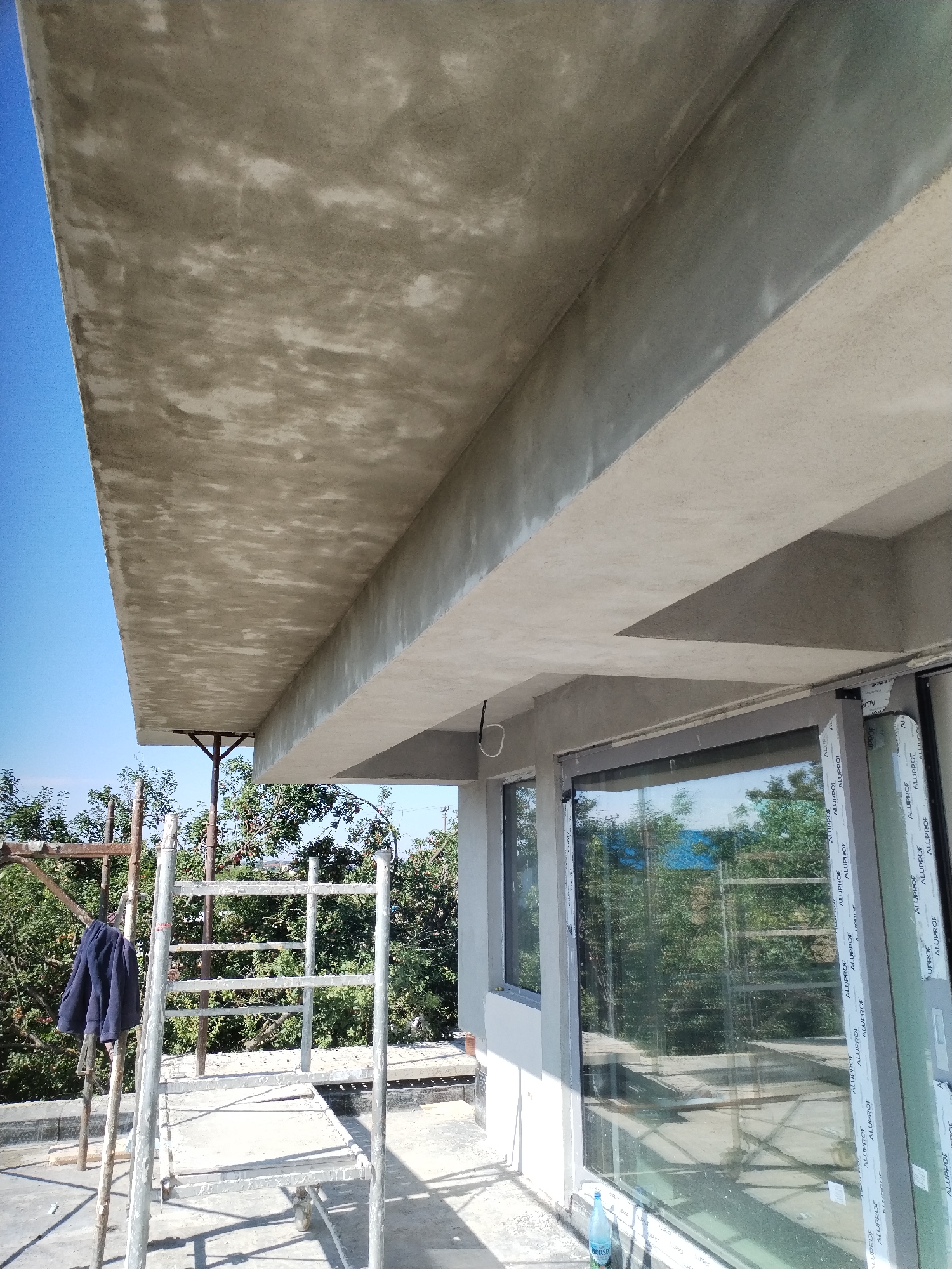 The image shows an unfinished concrete structure, likely a building's overhang or balcony, under a clear blue sky. The concrete surface appears rough and uneven, with visible variations in color and texture, indicating it is either freshly poured or has not yet been finished. Scaffolding is visible on the left side of the image, with a dark blue jacket hanging on one of the metal poles. The scaffolding suggests ongoing construction or renovation work. A large window or glass door is partially visible on the right, reflecting the surrounding trees and sky. The scene is set outdoors, with greenery and trees visible behind the structure and scaffolding. There are no people visible in the image. The overall impression is one of a construction site in an outdoor setting. The provided information indicates the location is in the city of Dumbrăveni, Romania.