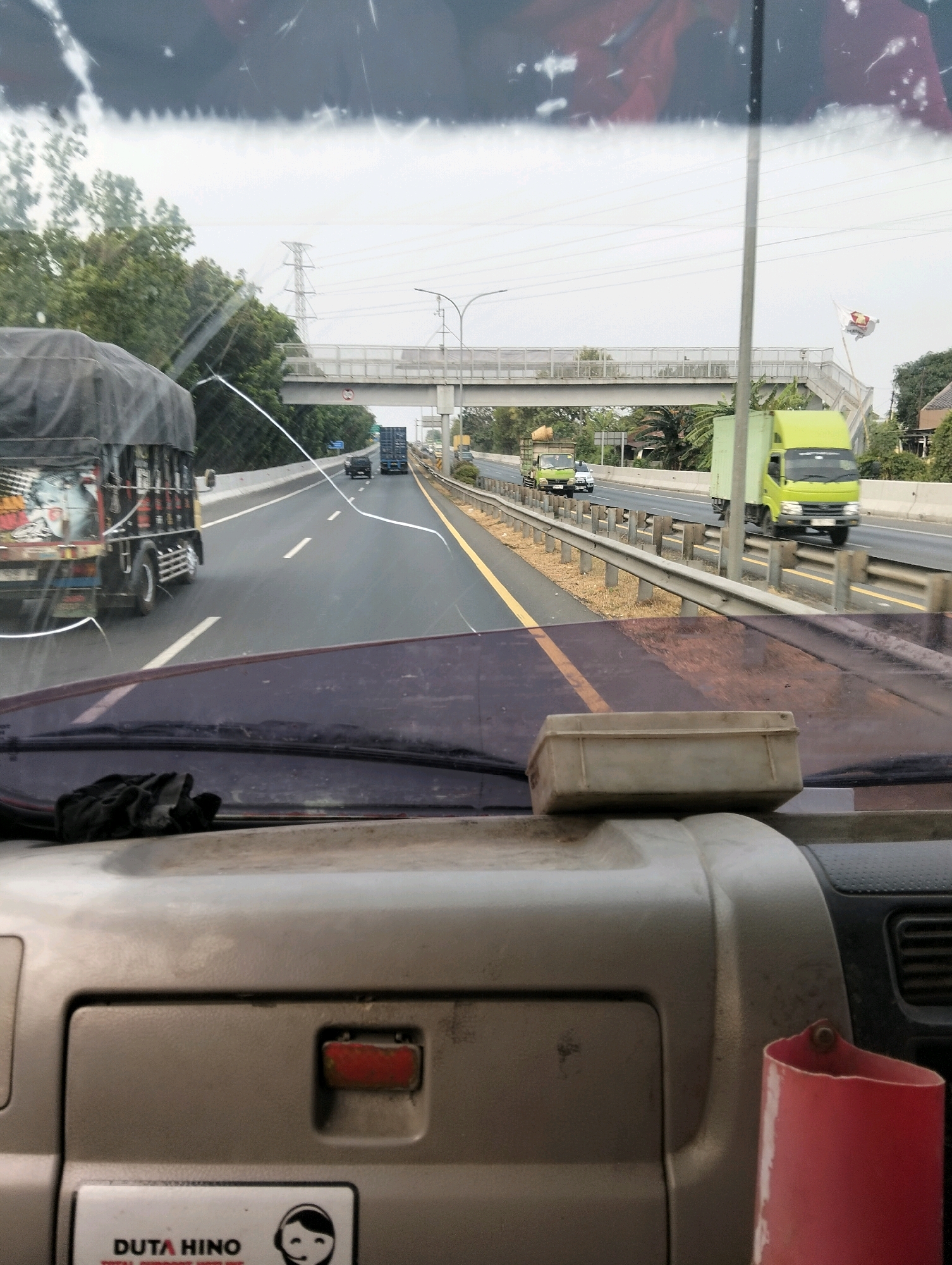 The image shows a view from inside a vehicle on a highway. The windshield has several streaks and smudges. In the foreground, on the dashboard, is a small, beige rectangular object. To the right of the object, a red cylindrical container is visible. A black cloth or rag is placed on the left side of the dashboard.
The highway is wet, suggesting it might have rained recently. Several trucks are visible on the road. A large black truck with a tarp-covered cargo is in the left lane. Further down the road, a blue shipping container truck is in the center lane. A bright green truck is in the right lane. In the distance, a pedestrian overpass spans the highway. There are trees and some buildings visible on the sides of the highway. Power lines and a transmission tower are also present in the background.
The text "DUTA HINO" and a logo are visible on a sticker in the lower left corner of the image, possibly indicating a dealership or service center related to Hino trucks, which are commonly used in Indonesia. While the image does not definitively place it in Semarang, Indonesia, the presence of Hino trucks and the style of highway infrastructure are consistent with locations in Indonesia, including cities like Semarang. There are no people clearly visible in the image. The overall scene depicts traffic on a highway under an overcast sky.
