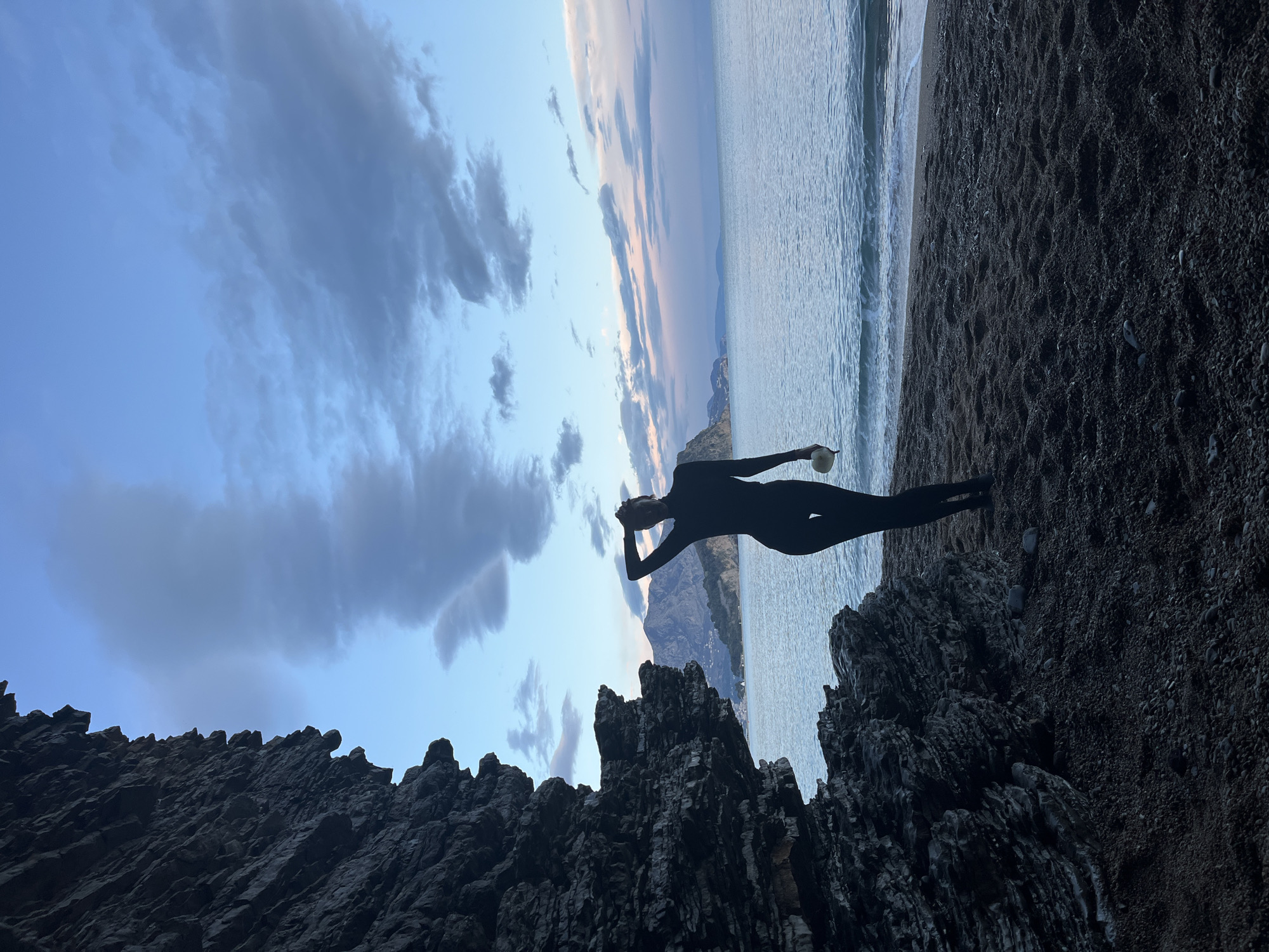 The image shows a silhouette of a person in a black, form-fitting suit standing on a rocky outcrop near the shore of what appears to be the Adriatic Sea. The person holds an object near their head and faces the sea. The background features a beach with dark sand, the sea with visible waves, mountains in the distance, and a sky with blue and grey clouds. The photo may have been taken in or near Budva, Montenegro, judging by the coastal landscape with mountains and rocky terrain.