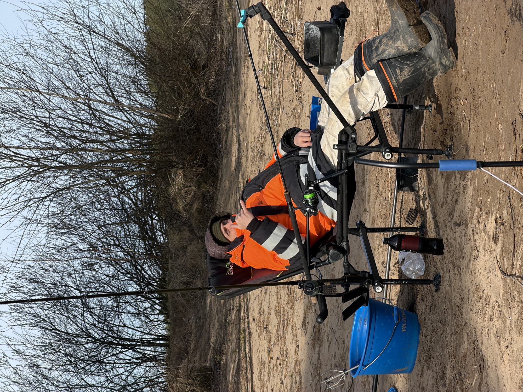 The image captures a man fishing on a sandy or dirt bank in Chaykovsky, Russia, likely during winter or early spring given the bare trees. The time of day is bright, sunny daylight.
The central subject is a man dressed in warm fishing attire, including an orange and black jacket, dark pants, a green beanie, and muddy black rubber boots with orange accents. He is seated comfortably in a specialized black fishing chair equipped with various attachments for rods and accessories. The man is looking towards the camera and making a "peace sign" gesture with his left hand, indicating a relaxed or positive mood.
Multiple fishing rods are visible, with one prominent rod extending across the foreground, fitted with a fishing reel. Another rod is held by a support attached to the fishing chair. Beside the chair are several items: a bright blue bucket with "VOLZIANO" printed on its side, a black rectangular container (possibly a bait box), a black bottle labeled "OLIVADO," and a clear plastic bottle. Part of a padded fishing bedchair or bag behind the man shows the text "FEEDER CONCEPT."
The setting features a wide expanse of light brown sandy or dirt ground. The background is filled with a dense mass of leafless trees and bushes against a clear, light blue sky. The overall scene suggests a calm outdoor fishing activity.