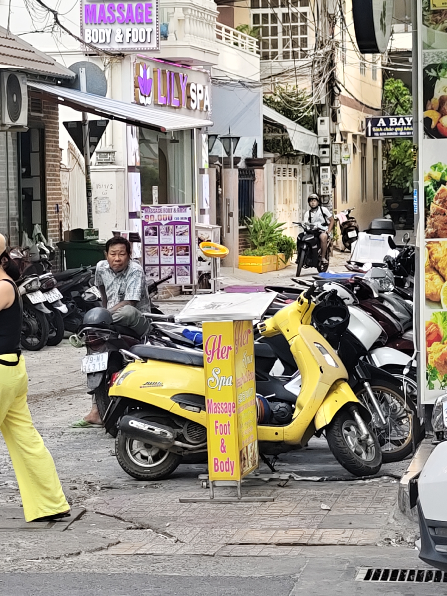 This image depicts a street scene in Nha Trang, Vietnam, likely during the daytime given the bright lighting. The foreground features a collection of parked motorcycles, dominated by a bright yellow scooter. A man in a checkered shirt sits on one of the motorcycles, looking towards the camera with a neutral expression. To his left, a person wearing bright yellow pants is partially visible, facing away from the viewer.
Behind the motorcycles, a row of businesses lines the street. A prominent sign above one establishment reads "MASSAGE BODY & FOOT" in large, illuminated purple letters. Below this, another sign for "LILY SPA" is visible. A smaller purple poster advertises "MASSAGE FOOT & BODY 90.000 / 30 MIN." Further down the street, a sign indicates "A BAY" with Vietnamese text.
The setting is a typical urban street with buildings, overhead electrical wires, and a tiled pavement. The presence of multiple massage parlors and the general street activity suggest a commercial district. The overall atmosphere appears to be one of everyday life, with the focus on the businesses and the vehicles that populate the street.