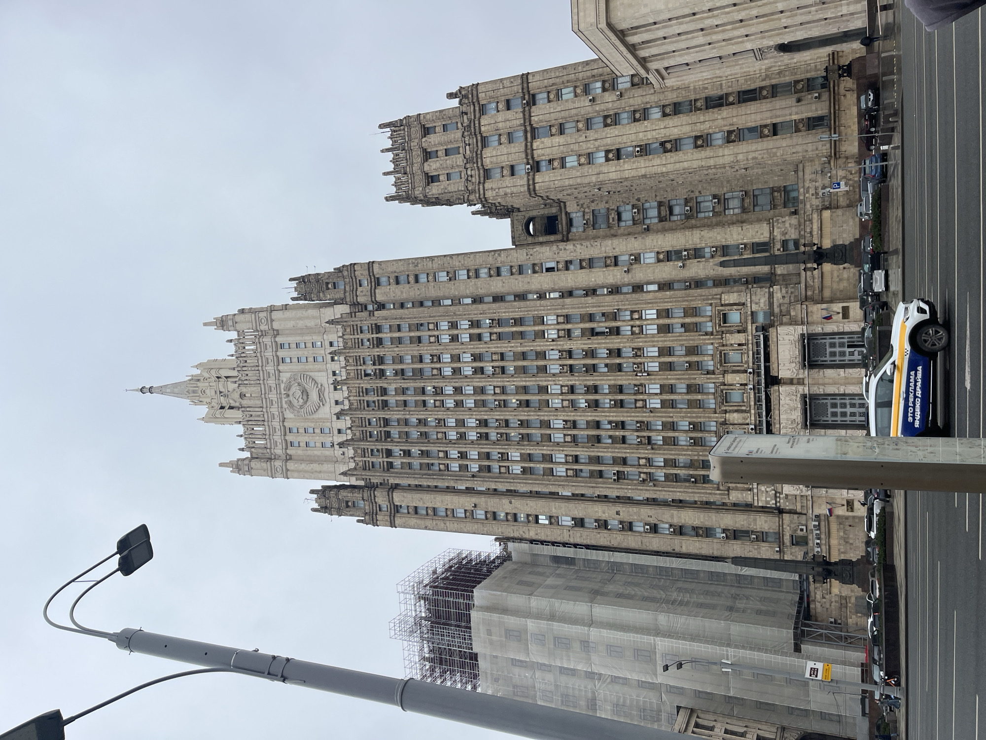 The image shows a tall, gray building in Moscow, Russia, under an overcast sky. The building has numerous windows and a prominent spire at the top, characteristic of Stalinist architecture. Scaffolding and a white shroud partially cover the lower portion of a nearby building, suggesting construction or renovation work. A streetlight pole is visible in the foreground on the left, and parked cars line the right side of the frame along a street. A vehicle with blue and yellow accents can be seen, suggesting a taxi or service vehicle. The composition appears to be taken from a low angle, emphasizing the height of the buildings. The scene appears to be an urban area with heavy vehicular traffic.