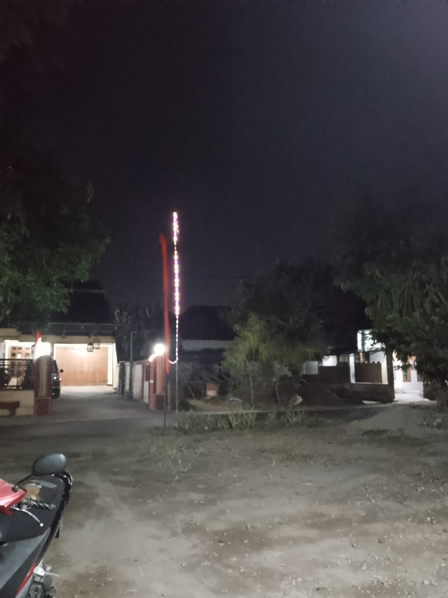 The image depicts an outdoor scene at night, likely in a rural or suburban area of Kelurahan, Indonesia, given the architecture and vegetation. In the foreground, the rear of a motorcycle is visible, with its black and red body. The ground is a mixture of dirt and gravel.
The mid-ground features a pathway leading to a building with a large open entrance, possibly a garage or carport. A car is partially visible within this entrance. To the right of the pathway, a red flag is mounted on a pole, accompanied by a string of decorative lights that run vertically up the pole, displaying multiple colors, likely pink or purple. Further to the right, more buildings with windows and roofs are visible, along with trees and foliage.
The background is dominated by the dark night sky. The overall impression is a quiet, residential setting at night, possibly decorated for an event or celebration.