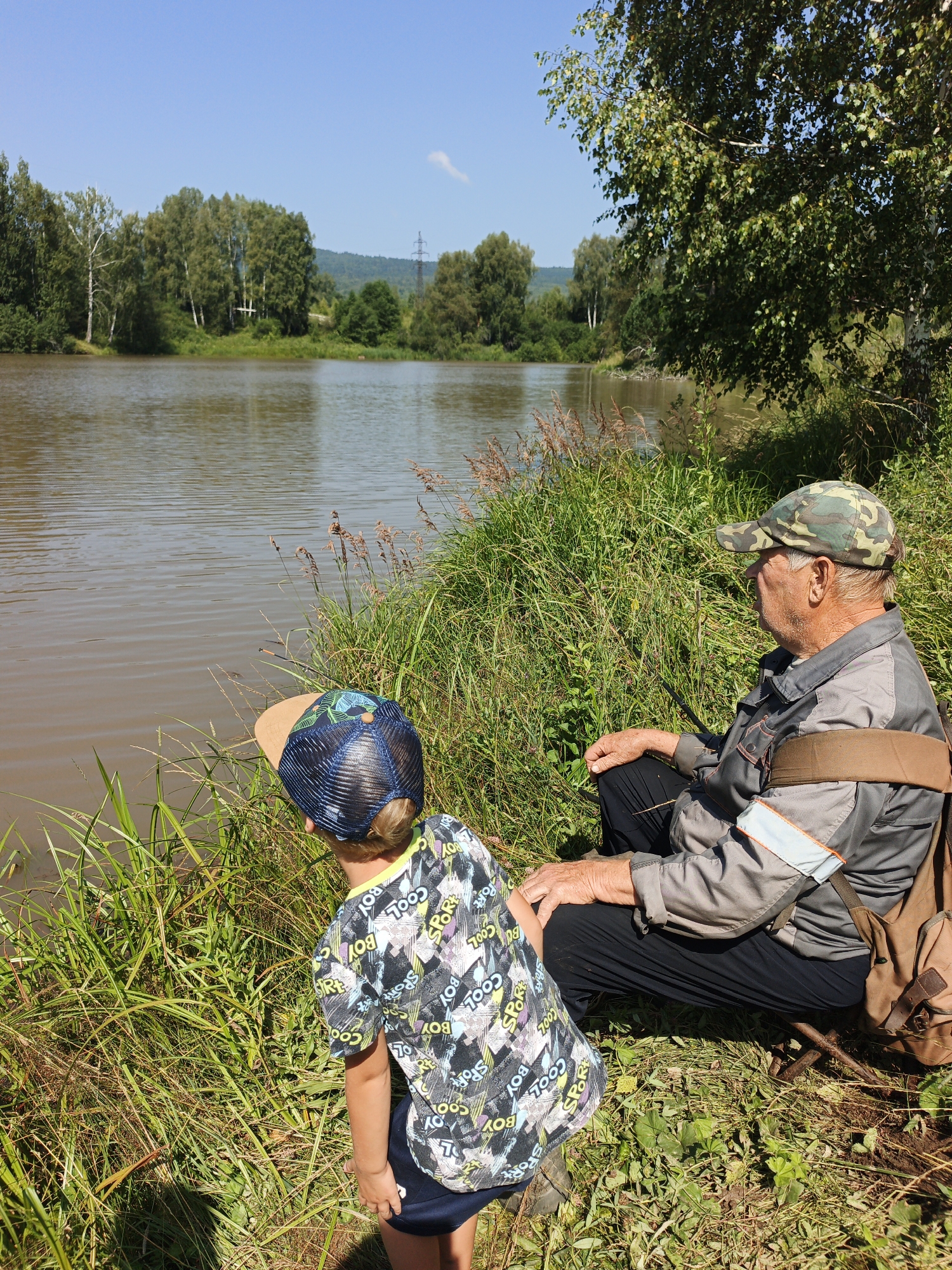 In Urtiushka, Russia, on a sunny day, an elderly man wearing a camouflage cap and a grey jacket sits by a river, looking out at the water. Beside him, a young boy in a patterned t-shirt and a blue baseball cap also faces the river, seemingly engaged in the same activity. The riverbank is overgrown with lush green grass and reeds. The background features trees and a distant hill under a clear blue sky.