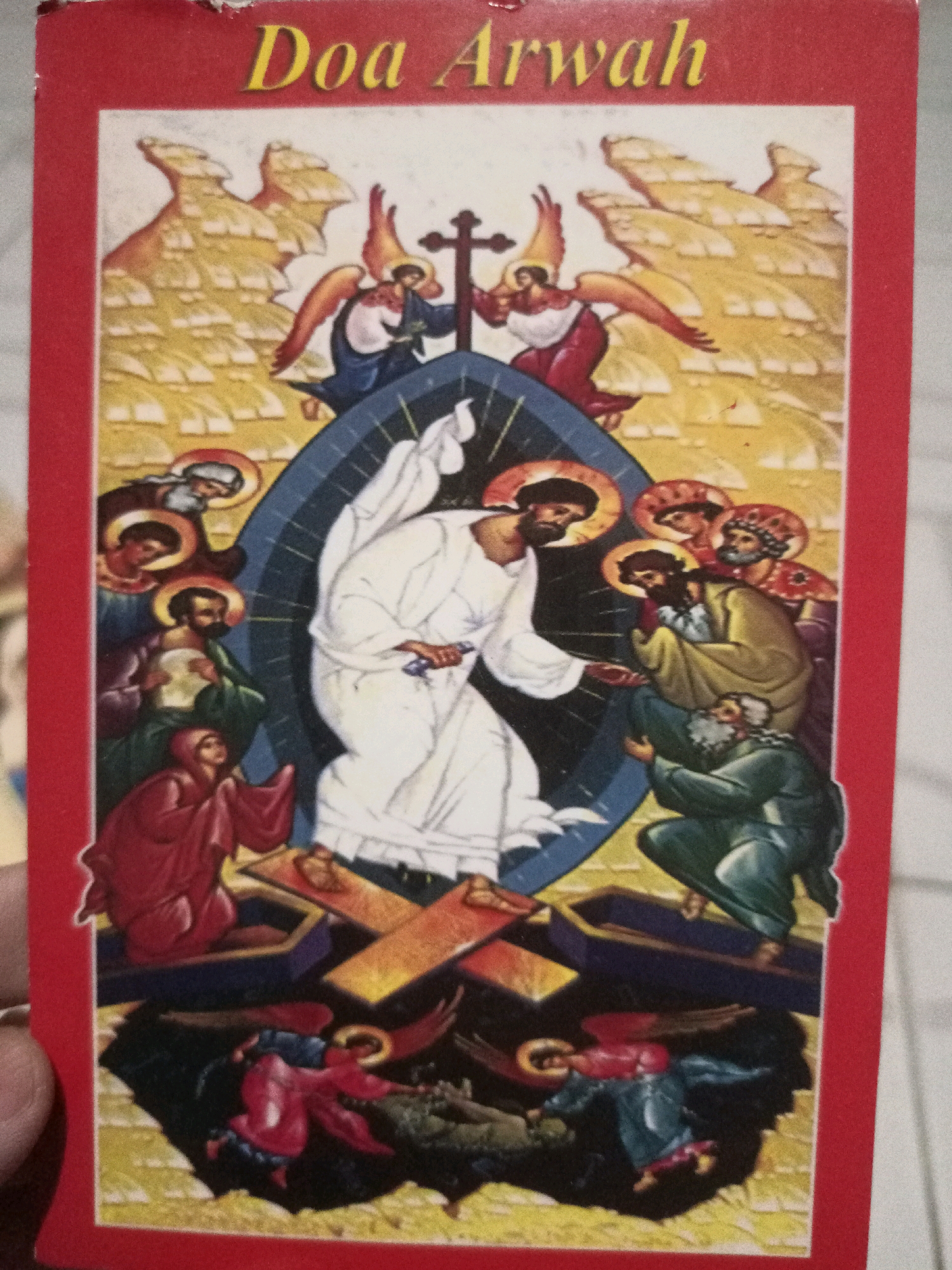 The image shows the cover of a book titled "Doa Arwah" in yellow text on a red border. The artwork depicts a religious scene, likely Christian, with a central figure in white robes, possibly Jesus, emerging from a blue oval shape. Around him are several figures with halos, appearing to be apostles or saints, dressed in various colored robes. Two angels with wings are positioned above and to the sides of the central figure, near a cross. Below, figures are shown emerging from open coffins, with other figures in the foreground, some appearing to be angels, tending to the scene. The background features abstract, rocky formations in shades of yellow and orange. The book cover is partially visible from the side, and the image is taken from a slightly downward angle, showing a white tiled surface beneath the book. There is no information available about the specific location in the city of Malang, Indonesia.