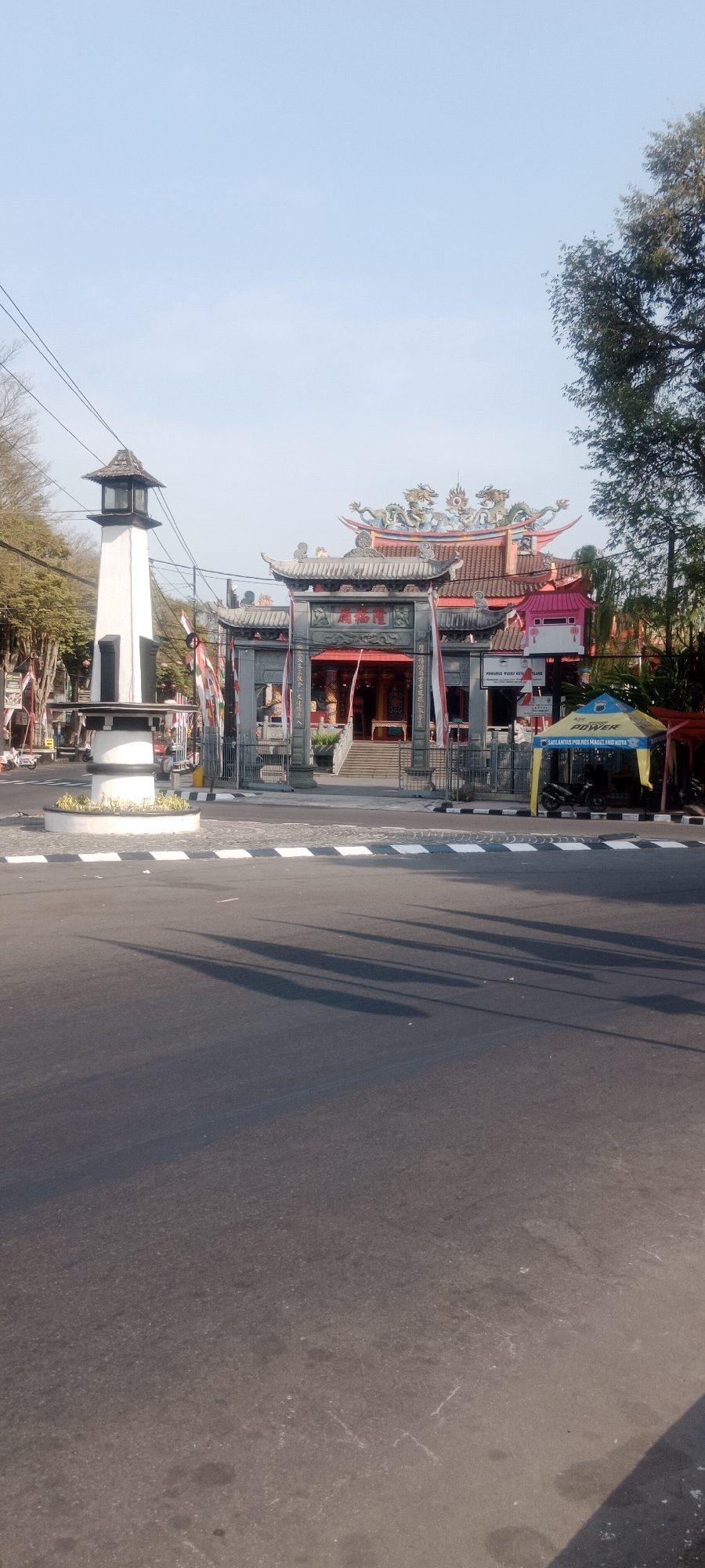 The image depicts a street scene in Magelang, Indonesia, featuring a prominent temple complex and a white tower. The temple, with its red roofs and ornate decorations including dragon motifs, is accessed through a large, traditional gate. In front of the temple, there are several flags and a blue and yellow tent. To the left of the temple, a tall, white, cylindrical tower with a lantern-like structure on top stands near a roundabout. The street in the foreground is paved and shows shadows cast by an unseen object. A few motorcycles are visible near the tent. The sky is clear and blue, and trees are present on the right side of the image.