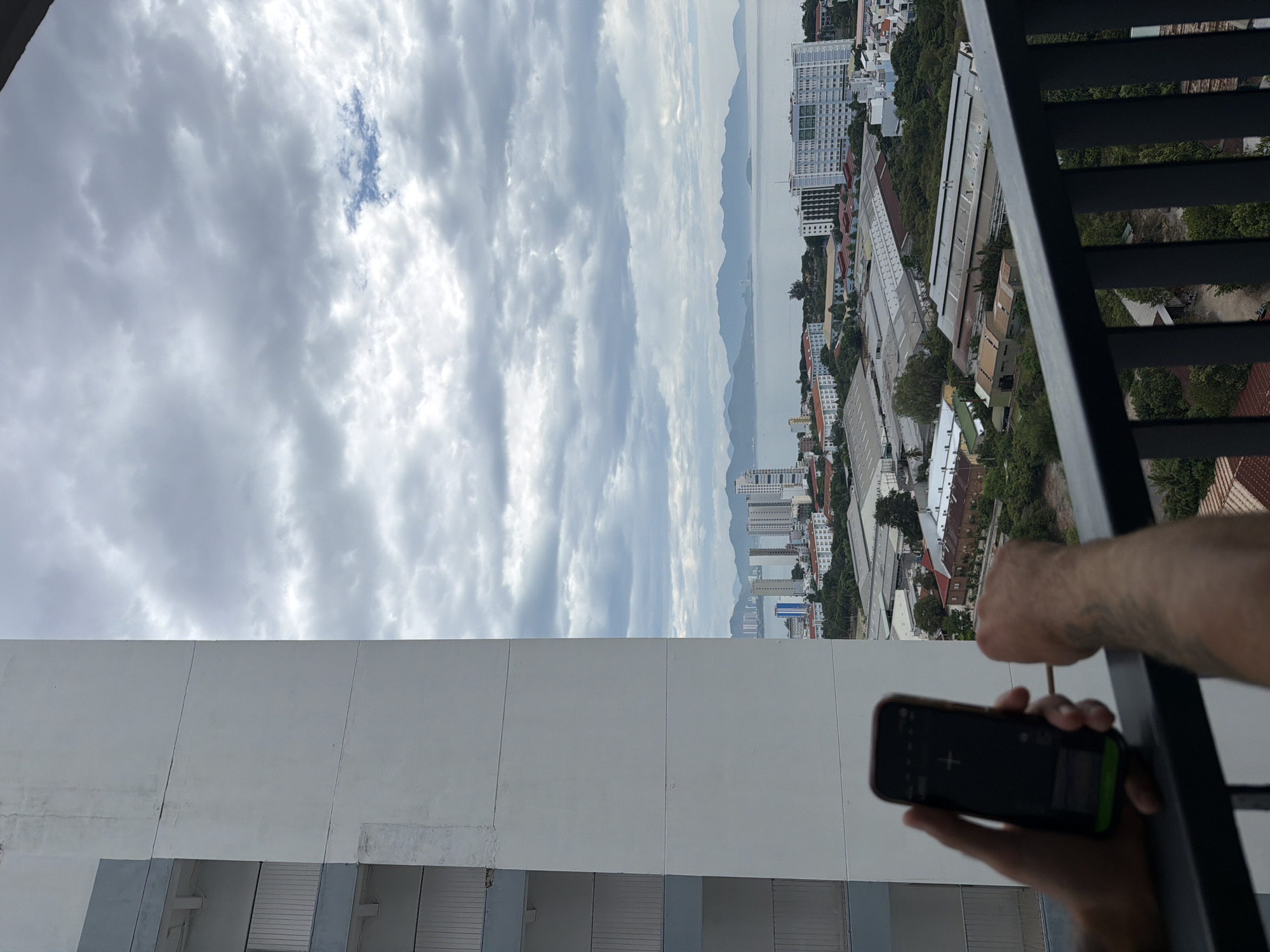 The image depicts a view from a balcony overlooking a city and coastline. In the foreground, a person's hands are visible holding a smartphone and what appears to be a slender stick or rod. The smartphone screen displays a dark interface with some illuminated elements. The balcony railing is dark metal.
The middle ground reveals a densely populated urban area with numerous buildings of varying sizes and architectural styles. Warehouses with corrugated metal roofs are prominent, interspersed with residential buildings and smaller structures. Greenery, including trees, is scattered throughout the urban landscape.
The background features a wide expanse of the sea, with a distant coastline characterized by rolling hills and mountains under a cloudy sky. The sky itself is filled with a mix of dark and lighter gray clouds, with a small patch of blue sky visible. The overall lighting suggests an overcast day. The architecture and the presence of a bay suggest the city of Nha Trang, Vietnam.