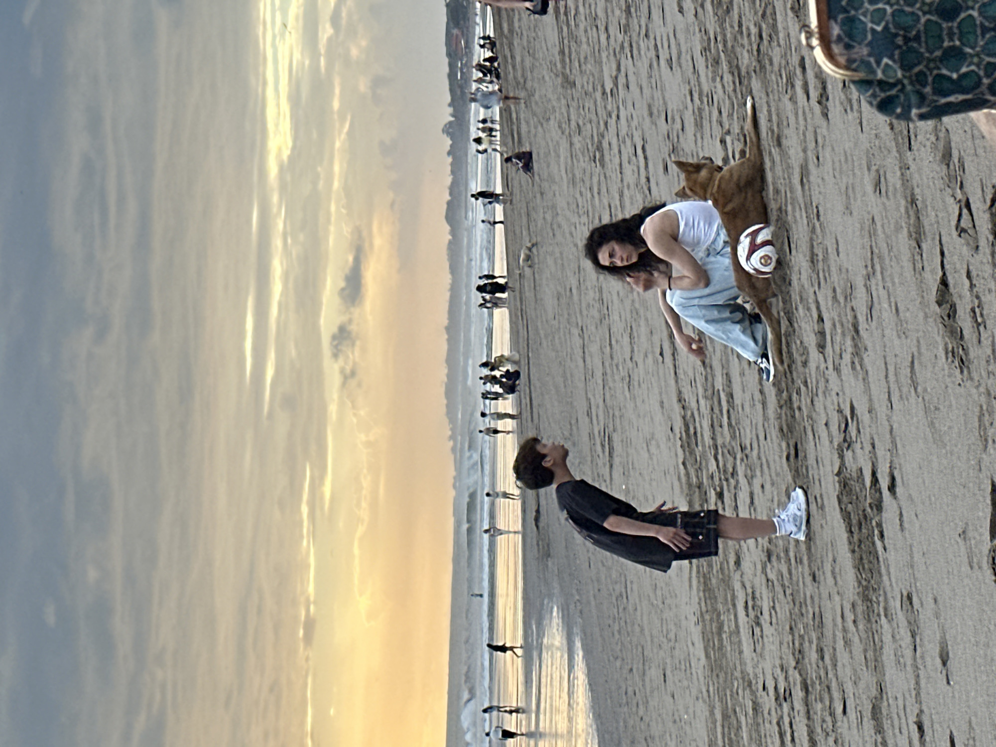 This image depicts a beach scene in North Kuta, Indonesia, during what appears to be sunset. In the foreground, a young boy wearing a black t-shirt and shorts stands facing a young woman. The boy is looking up at the woman, with his hands extended. The woman is seated on the sand, wearing a white tank top and light blue pants. She is looking off to the side, and her left hand is raised to her mouth. Next to the woman, a brown dog lies on the sand with a soccer ball positioned between its hind legs. The beach is populated with other people in the background, many of whom are walking along the water's edge. The ocean has moderate waves, and the sky is filled with clouds, with a warm yellow and orange glow indicating the setting sun. The sand on the beach has a textured appearance with visible footprints and debris.