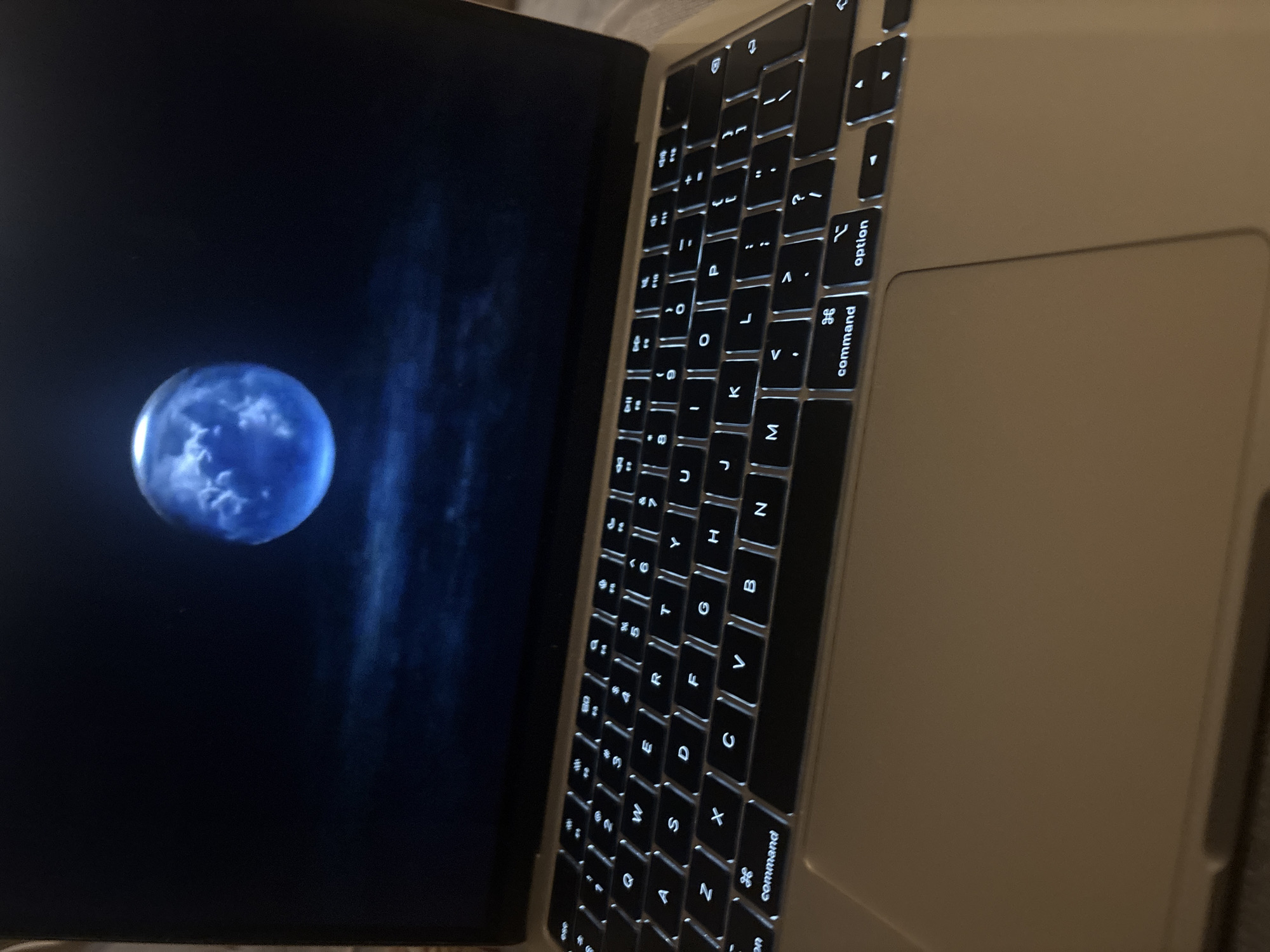 An illuminated laptop keyboard is in focus, with the screen displaying a blue moon against a dark, cloudy sky. The laptop is positioned on a light-colored surface, possibly a desk or table. The keyboard keys are backlit, and several keys, including "command" and "option," are clearly visible. The scene appears to be indoors in a dimly lit environment. There are no people visible in the image. The location is not specified within the city of Sofia, Bulgaria.