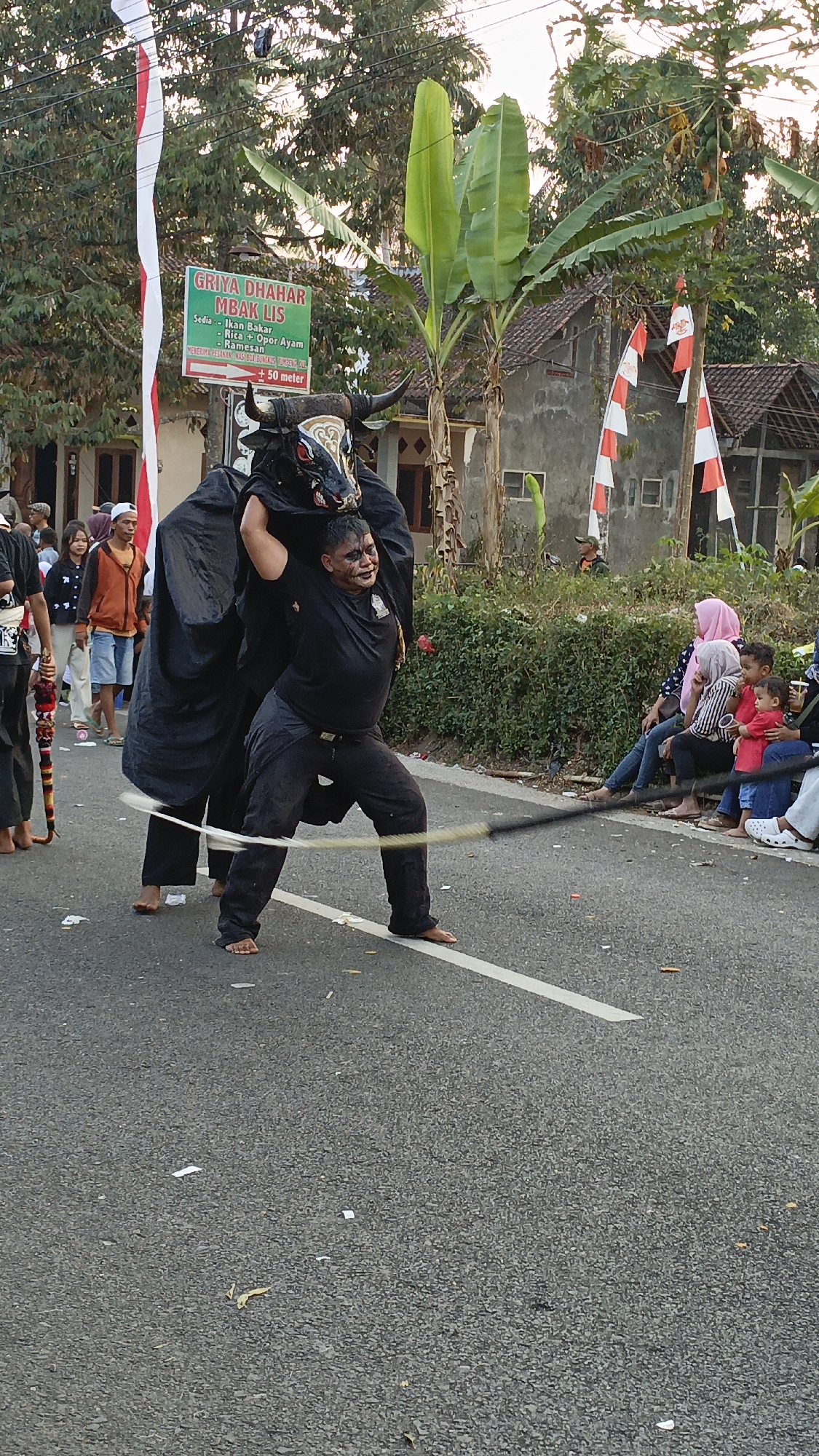 In the city of Krajan Lor, Indonesia, a cultural performance is underway on an asphalt road. Two individuals are engaged in a reenactment of a bull, with one person wearing a bull mask and costume, and the other person supporting the costume from behind. The person in front, dressed in black, appears to be the main performer, holding a whip-like object and exhibiting an energetic stance. They have theatrical makeup on their face, suggesting a dramatic portrayal. Spectators are gathered on the side of the road, with some seated on a low wall and others standing. The environment includes residential buildings, lush green vegetation, and decorative Indonesian flags. The presence of a signpost in the background indicates a commercial establishment, adding to the local context of the scene. The overall atmosphere suggests a vibrant community event or festival.