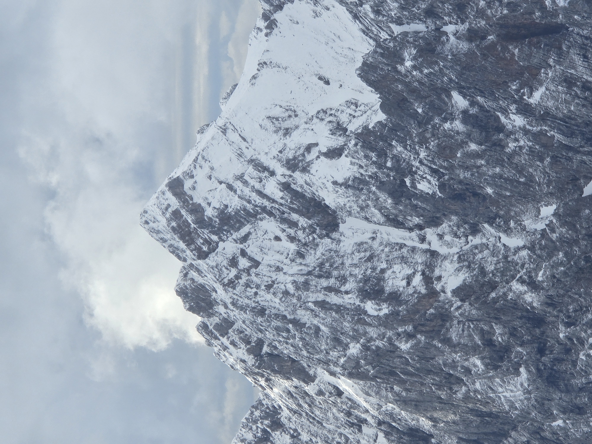 The image shows a snow-covered mountain peak. The mountain has a rugged, rocky texture, with patches of snow clinging to its slopes and a thin dusting of snow on its summit. The sky above is overcast with scattered clouds, some of which are illuminated by sunlight. The scene is likely part of the Bavarian Alps, which surround the city of Garmisch-Partenkirchen, Germany. There are no people or man-made objects visible in the image. The overall impression is one of a majestic and remote natural landscape.