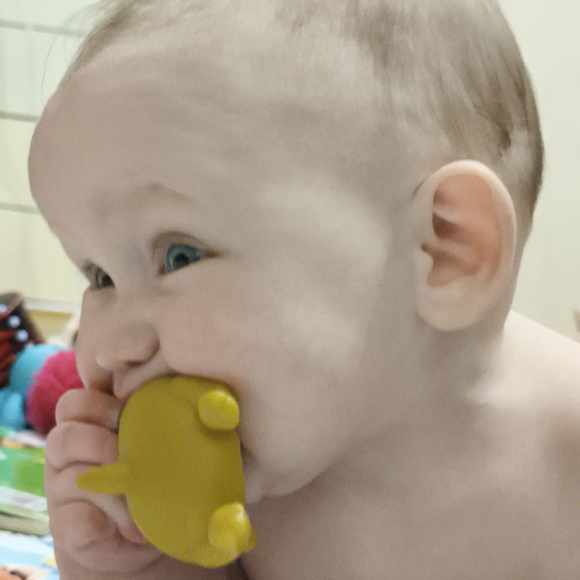 A baby with light skin and blonde hair is shown in a close-up, side-profile view. The baby is holding a yellow, animal-shaped toy in their left hand and has part of it in their mouth. The baby has blue eyes that are looking off to the side. The background includes blurred colorful objects, suggesting a play area. The location and any information about the city of Chaikovsky, Russia, is not directly evident from the image.