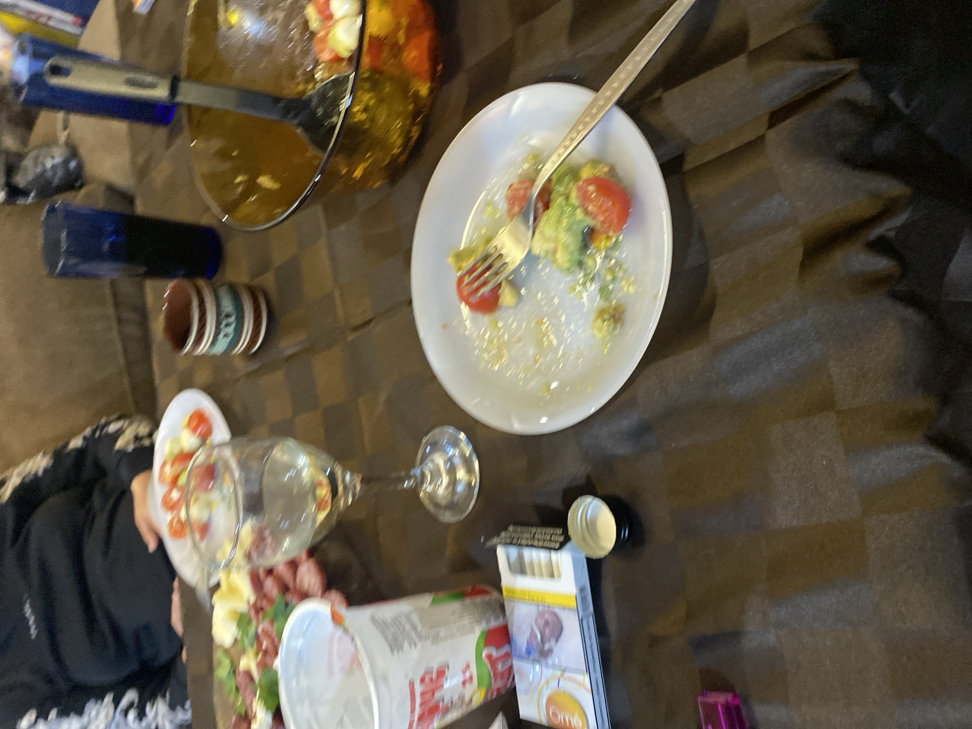The image shows a cluttered tabletop scene, possibly indoors. A partially eaten salad plate with a fork is in the center. A bowl with salad and a serving spoon is to the upper left. Other items include a wine glass, various food containers (one labeled "Oma's"), a pack of cigarettes, and various small objects. Part of a person wearing a black and white patterned garment is visible on the left. The location is not discernible from the image; there is no information in the image that indicates it is located in Belopoptsi, Bulgaria.