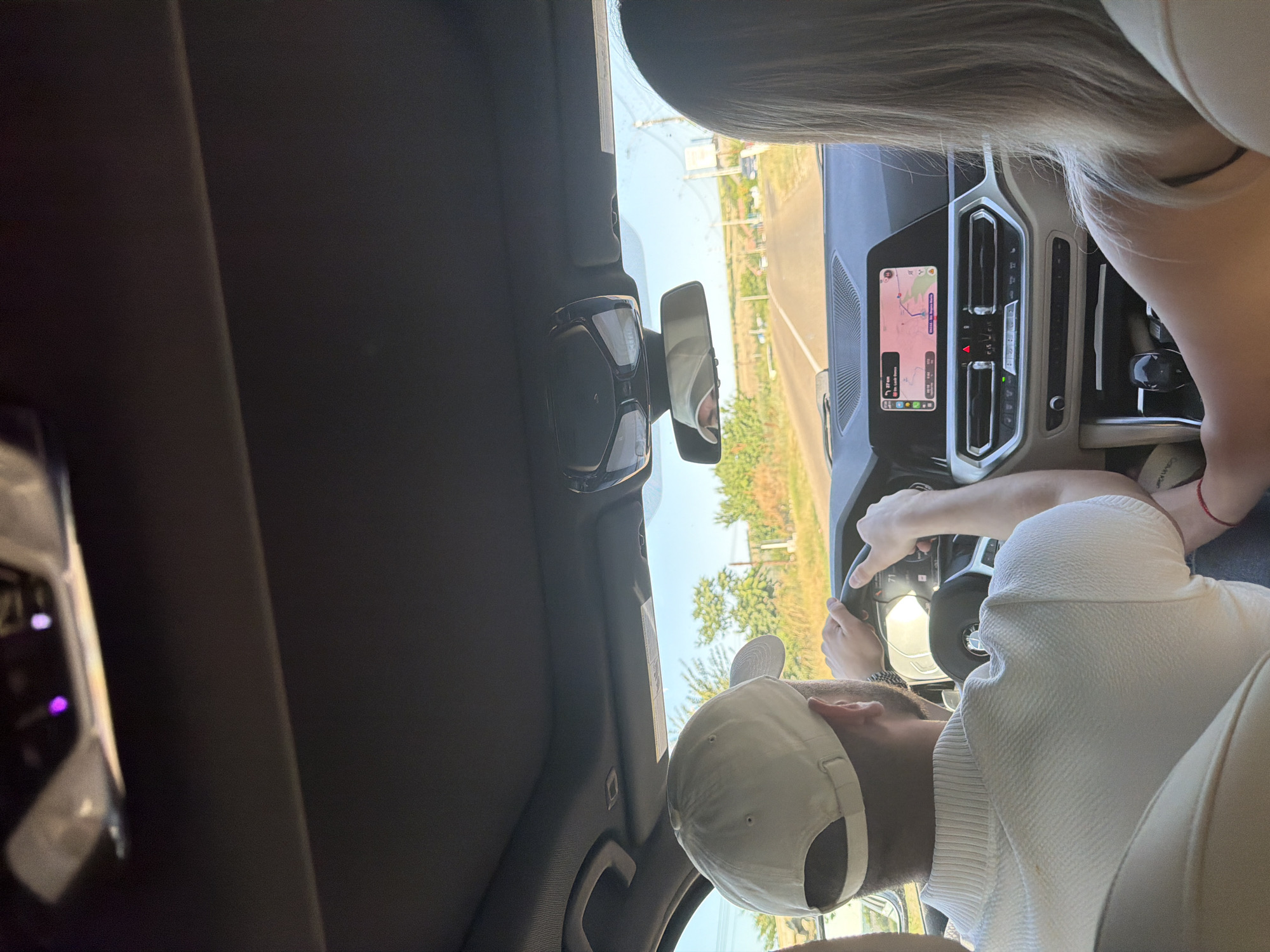 The image shows the interior of a car, taken from the perspective of a passenger. A man wearing a white cap and a white collared shirt is driving. He is holding the steering wheel with both hands, and his back is to the camera. A woman, also with her back to the camera, is sitting in the passenger seat. Her blonde hair is visible. A red bracelet is visible on her wrist. The car's dashboard and navigation screen are visible. The navigation screen shows a map with the route to Zorleni, Romania. Outside the car, trees and a road are visible. The sky is blue.