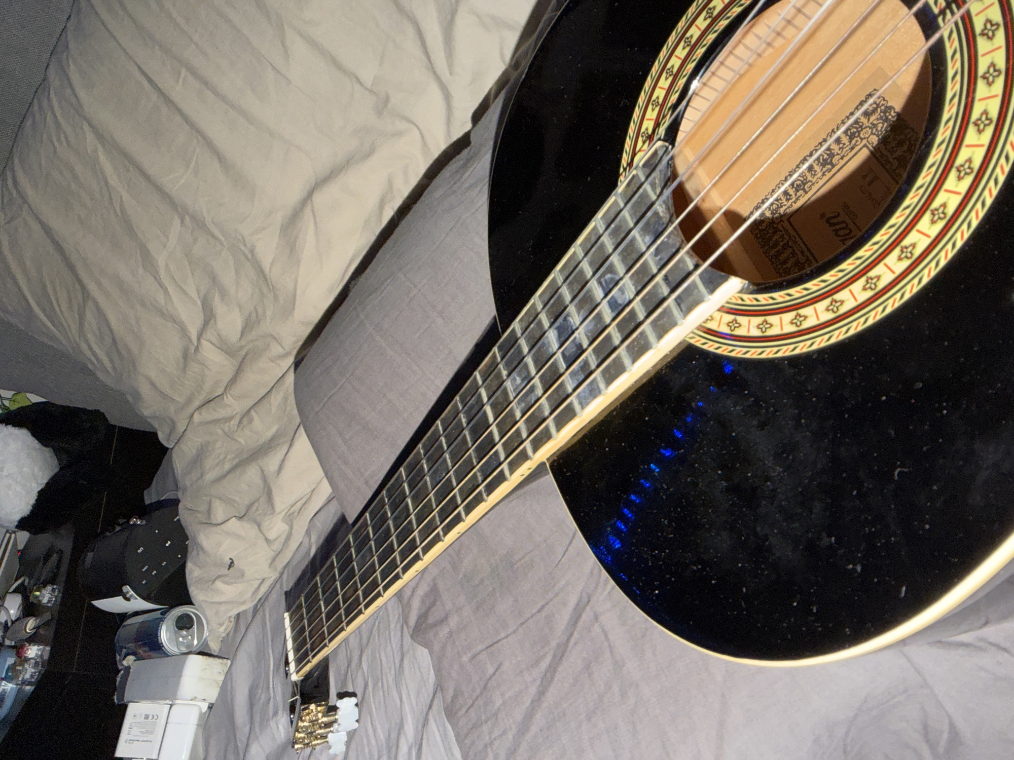 The image shows a black acoustic guitar lying on a bed. The guitar is positioned diagonally across the frame, with the neck and fretboard extending towards the top left corner. The body of the guitar is in the foreground, and its soundhole is visible, revealing some text on a label inside. The guitar has nylon strings and gold-colored tuning pegs. The bed is covered with grey and beige linens, and a hint of a pillow can be seen in the background. On a surface to the left of the bed, there are various objects, including a white power adapter, a blue and silver can, and a black electronic device. The lighting is dim, with some blue light reflections visible on the guitar's body. The image does not provide any information about the location within the city of Oslo, Norway.
