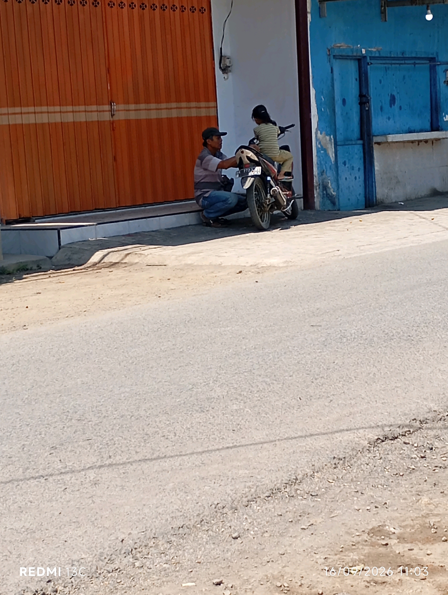 An adult male wearing a striped shirt and a dark baseball cap is seated on the ground next to a motorcycle in front of a large, orange, corrugated metal shuttered storefront. A young person, possibly a child, wearing a striped shirt and tan pants is seated on the back of the motorcycle. The scene appears to be outdoors on a paved surface, possibly a street or sidewalk, with a road in the foreground. The background shows a blue building with a closed door and a white wall. The timestamp "16/09/2026 11:03" is visible in the lower right corner of the image, and "REDMI 13C" is in the lower left. The location is identified as Wuluhan, Indonesia.