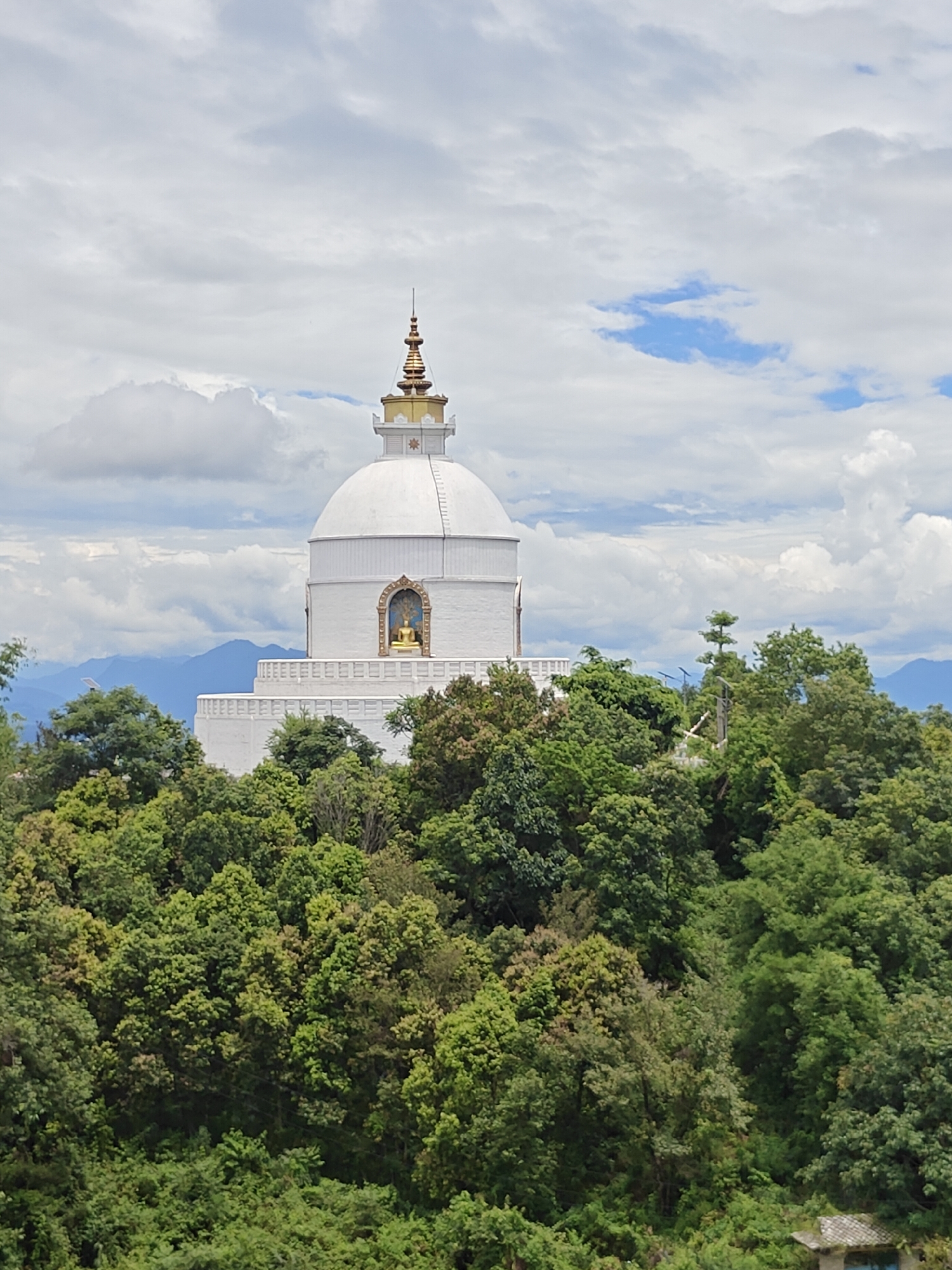 The image shows the World Peace Pagoda in Pokhara, Nepal. The pagoda is a large white dome-shaped structure with a golden spire on top, situated on a hilltop surrounded by lush green trees. The sky is partly cloudy with patches of blue sky visible. In the distance, hazy blue mountains can be seen. The pagoda features a statue of Buddha in an alcove on its side. The overall scene conveys a sense of serenity and natural beauty.