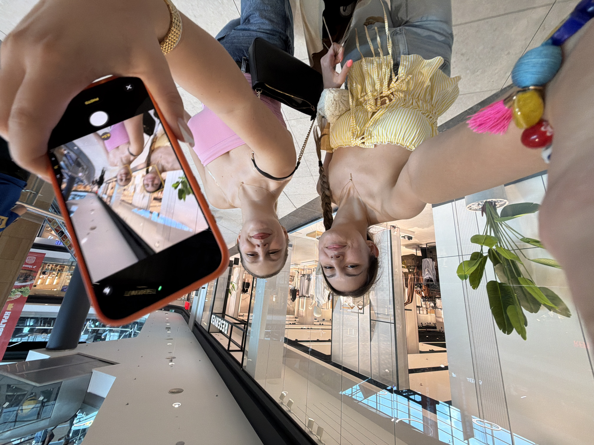 The image captures two young women and parts of a third person in what appears to be a modern shopping mall in Burgas, Bulgaria. The primary subjects, the two women, are presented in an inverted perspective, strongly suggesting they are reflections captured from a highly polished floor or a similar reflective surface.
One woman, positioned to the left, wears a light pink sleeveless crop top and blue jeans, with a black shoulder bag slung over her. Her hand, adorned with a gold-colored bracelet, holds a smartphone with an orange case. The phone's screen displays a live camera view, showing the reflections of both women, appearing right-side up, with "PHOTO" and the shutter button visible on the interface. The second woman, on the right, wears a yellow and white striped crop top and light blue jeans. She has a colorful beaded bracelet with a vibrant pink tassel on her wrist and her hair in a braid. Both women are smiling, indicating a cheerful and relaxed mood.
The background, also appearing inverted due to the reflective surface, reveals typical mall architecture: high ceilings, glass shopfronts, and bright, diffused indoor lighting. Notable details in the background include a potted plant with large green leaves. Visible text on reflected shop signs includes "RESERVED" and parts of a "BURGER KING" logo, along with Cyrillic text such as "KPAЛ...". The image captures an activity of taking a selfie or a photo of reflections within the mall environment.