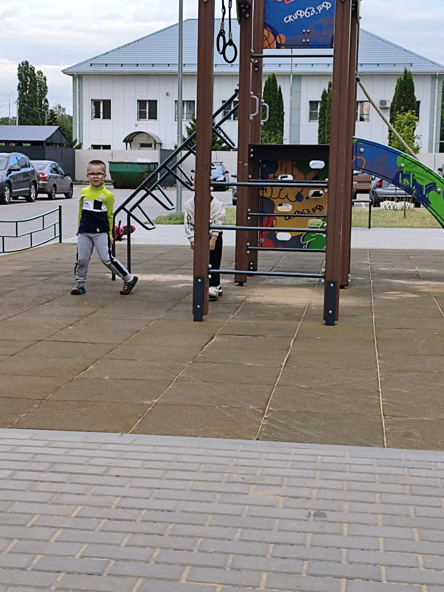 This media file shows an outdoor scene at a playground. In the foreground, a young boy with glasses and a lime green and black long-sleeved shirt, grey pants, and black shoes stands facing left. He is positioned on a rubberized tiled surface. Behind him, another child, mostly obscured, is seen from the waist down, wearing a white top and black pants.
The playground equipment consists of a large wooden structure with metal bars and climbing elements, including what appears to be a climbing wall with colorful graphics. A green slide is partially visible on the right.
In the background, a modern white building with a grey roof is situated. There are parked cars, including a dark grey sedan and a silver sedan, on a paved area to the left of the playground. A large green dumpster is also visible near the building. The ground beyond the playground is covered in grass and more paving.
The sky is overcast, suggesting cloudy weather. The lighting indicates it is daytime, possibly late afternoon or early evening. There is no specific indication of the location or any particular event taking place, beyond children playing.
Visible text on the playground equipment includes "скиф63.рф" in blue lettering on a colorful panel at the top, and a similar text appears on a lower panel.