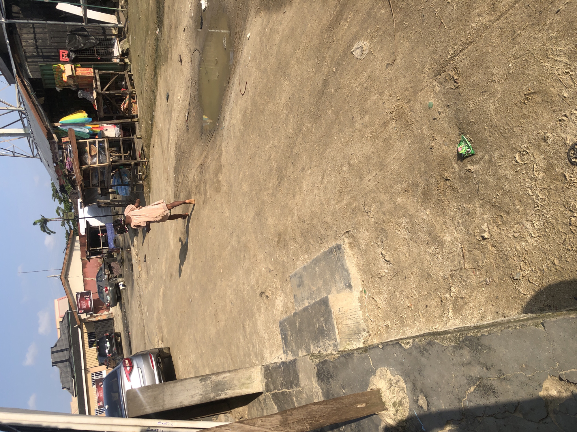An image taken in Ado, Nigeria depicts a dusty street scene. A young girl wearing a light-colored dress is walking away from the camera, her back to the viewer, on the right side of the frame. Her arms are outstretched as she walks, suggesting she might be playing or running. Her shadow is cast on the ground. In the background, several vehicles are parked, including a silver sedan on the left and a dark SUV further back. Buildings with corrugated metal roofs and some commercial stalls are visible behind the vehicles. A small puddle of water is present on the ground in the mid-ground. The overall environment is characterized by unpaved ground and a somewhat disorganized collection of structures, indicative of a developing urban area. The sky is clear and blue, suggesting a sunny day.