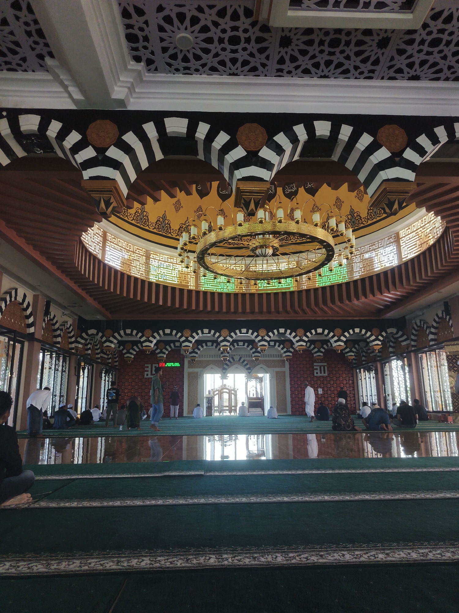 The image shows the interior of a mosque in Beringin, Indonesia. The perspective is from the floor, looking towards the front of the prayer hall. The floor is covered with green carpet, with patterned borders on some sections. Several people are seated and kneeling on the carpet, some facing forward, suggesting prayer or reflection.
The architecture of the mosque is ornate, with a large, circular chandelier hanging from the high, domed ceiling. The ceiling itself is decorated with intricate geometric patterns and calligraphy. Arches with black and white striped detailing are visible, supporting the upper levels of the structure. Wooden beams are incorporated into the ceiling design, adding warmth.
Light streams in from windows and openings, illuminating the space and casting reflections on the polished floor in the mid-ground. The walls are decorated with red patterned tiles and calligraphy. A digital display showing a time of 12:10 is visible on one of the walls, alongside the word "ISLAM". The overall impression is one of reverence and architectural beauty within a place of worship.