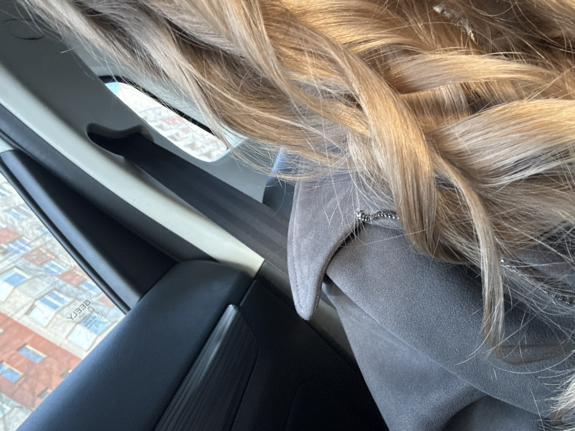 The image depicts a close-up view from the interior of a vehicle, likely the front passenger seat. In the foreground, the upper back and right shoulder of an individual are visible, adorned in a textured, grey or light brown garment, possibly a coat or jacket, featuring a silver-colored chain detail near the shoulder. A significant portion of long, wavy, blonde hair, illuminated by direct sunlight, flows across the lower right quadrant of the frame. A black seatbelt is discernible across the person's shoulder area.
The vehicle's dark grey interior paneling is visible on the left, while a lighter, possibly beige, pillar frames the upper right. Through the vehicle's window, an urban street scene is reflected and viewed. The exterior environment consists of multi-story residential buildings: one predominantly red brick structure with regularly spaced windows, and another lighter grey, possibly concrete or stucco building with similar window patterns, some reflecting a blue sky. Patches of clear blue sky are visible between and above the buildings. Reflected on the window glass is inverted Cyrillic text, clearly reading "БЕГГА" (BEGGA), alongside a circular emblem, suggesting external signage. The architecture and the visible Cyrillic text firmly place the scene in an urban residential area within Saint Petersburg, Russia. The precise street or district cannot be determined from the available visual information beyond its general character as a typical city residential environment.