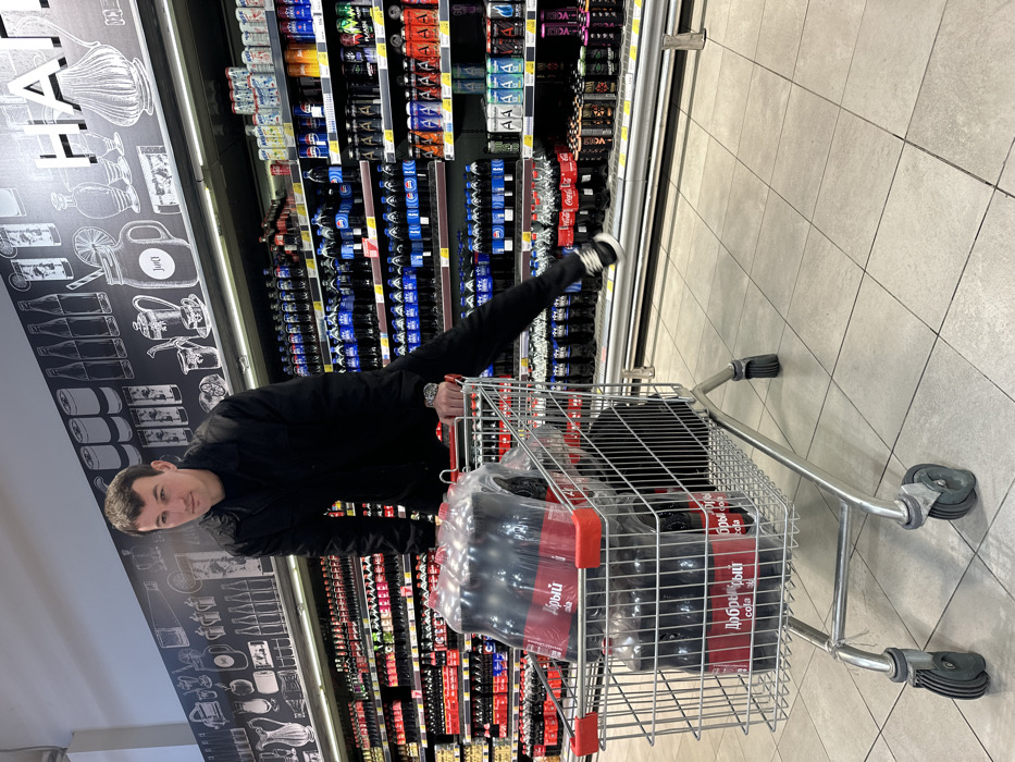 An adult male, approximately 25-35 years old, is depicted in the beverage aisle of a supermarket in Sochi, Russia. He wears a black long-sleeved shirt or light jacket, black pants, and black sneakers with white soles. A silver-toned watch is visible on his left wrist. He is positioned with his torso leaning over a silver metal shopping cart, his left leg extended horizontally to the right, elevated above the lower shelves. His right hand grips the shopping cart's red plastic handle. He maintains a neutral expression with a slight smirk, looking directly forward. The shopping cart contains at least three multi-packs of "Добрый Cola" (Dobry Cola), consisting of multiple dark-colored bottles with red labels, wrapped in clear plastic.
The background shows two fully stocked beverage shelves extending into the distance. The upper shelf features numerous rows of dark-colored soft drink bottles, including identifiable brands like Coca-Cola and Pepsi, along with various blue-labeled alternatives. The lower shelf displays an assortment of canned beverages, including energy drinks such as Flash Up, and other colorful soft drinks. Above the shelves, a dark wall panel is decorated with white chalk-style illustrations of bottles, glasses, and drink concepts. Partially visible Russian text on this panel includes "НАПИТКИ" (Beverages). The floor is composed of light-colored square tiles. The setting is brightly lit, consistent with an indoor retail environment.
