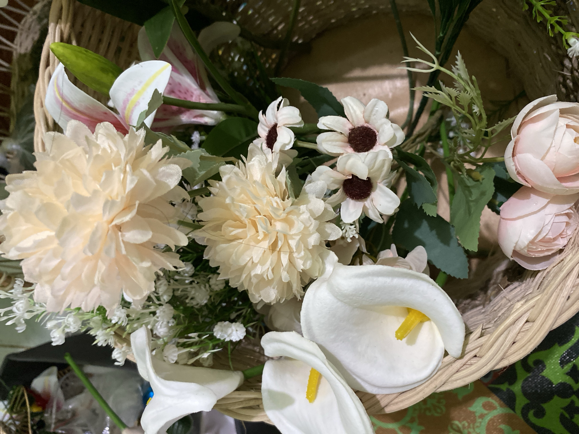 This media file is a close-up, overhead shot of a decorative arrangement of artificial flowers in a woven basket. The subjects are the various types of flowers, predominantly in shades of cream, white, and pale pink, along with green foliage. Some of the identifiable flowers include large, fluffy blooms that resemble peonies or chrysanthemums, smaller daisy-like flowers with dark centers, and elegant white calla lilies with bright yellow spadices. There are also hints of pink and white lilies, possibly Stargazer lilies, in the background. The setting is a basket with a rustic, woven texture, suggesting it could be used for display or as part of a gift arrangement.
There is no discernible activity or event taking place; the scene is static, focusing on the visual composition of the flowers. The lighting appears to be artificial and somewhat soft, casting subtle shadows within the basket and among the petals. There are no clear cues to indicate the time of day or weather conditions. The location context provided places this scene in Kuala Pembuang, Indonesia, but there are no explicit visual elements within the image itself that directly confirm this location, such as specific architectural styles or local flora beyond the artificial arrangement.
Notable details include the variety of textures and shapes of the flowers, from the delicate petals of the smaller blooms to the substantial forms of the calla lilies and the fluffy blooms. The basket's weave is also a prominent textural element. The overall aesthetic is one of floral beauty and arrangement. No text is visible in the image.