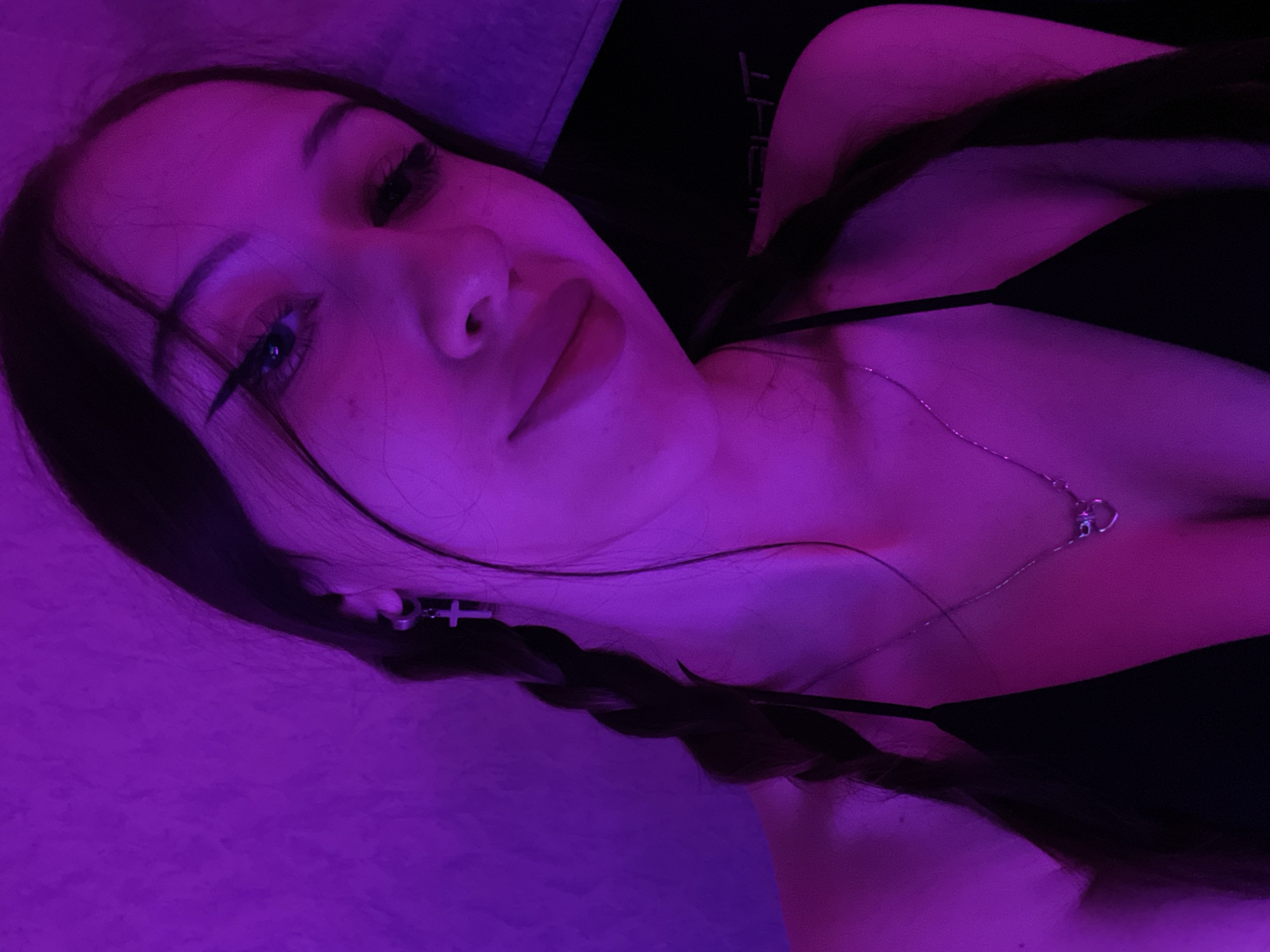The image is a close-up selfie of a young woman indoors, illuminated by purple lighting. She has her hair in two braids and is wearing a black top and a necklace with a heart pendant. She is also wearing a cross earring. The background appears to be a textured wall and a dark surface. There is no visible indication of the city of Votkinsk, Russia, in the image.