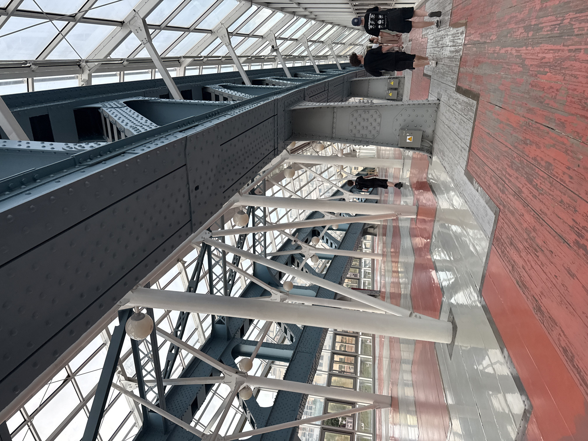 The image shows the interior of the Bridge of the Seven Bridges, a pedestrian bridge in Moscow, Russia. The bridge is enclosed in glass and features a complex steel structure with blue and white beams. Several people are walking along the red and gray wooden floor. The lighting is natural, coming from the glass ceiling.