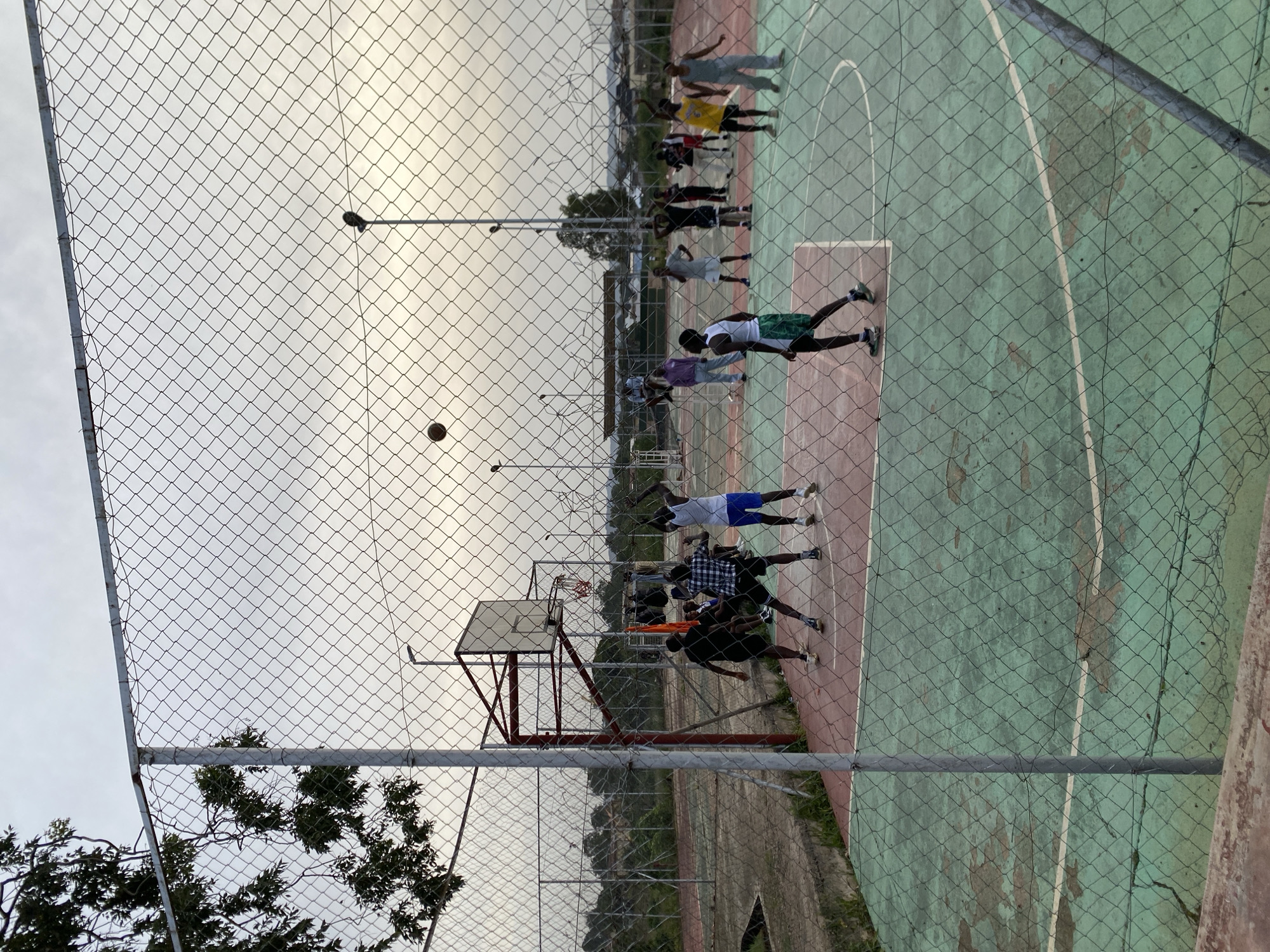 The image depicts an outdoor basketball court with a chain-link fence in the foreground, partially obscuring the view. A group of young men are engaged in a basketball game on the court. The court surface is green with red accents and appears worn in places. A basketball is visible in the air. Several players are in motion, with one player in a white tank top and blue shorts jumping to shoot, another in a checkered shirt and dark shorts is defending, and a player in a white tank top and green shorts is also actively participating. In the background, more individuals are watching or standing around the court, and there are trees and buildings visible in the distance under an overcast sky. The setting is consistent with a recreational area in the city of Jos, Nigeria, likely a public park or a school's sports facility.