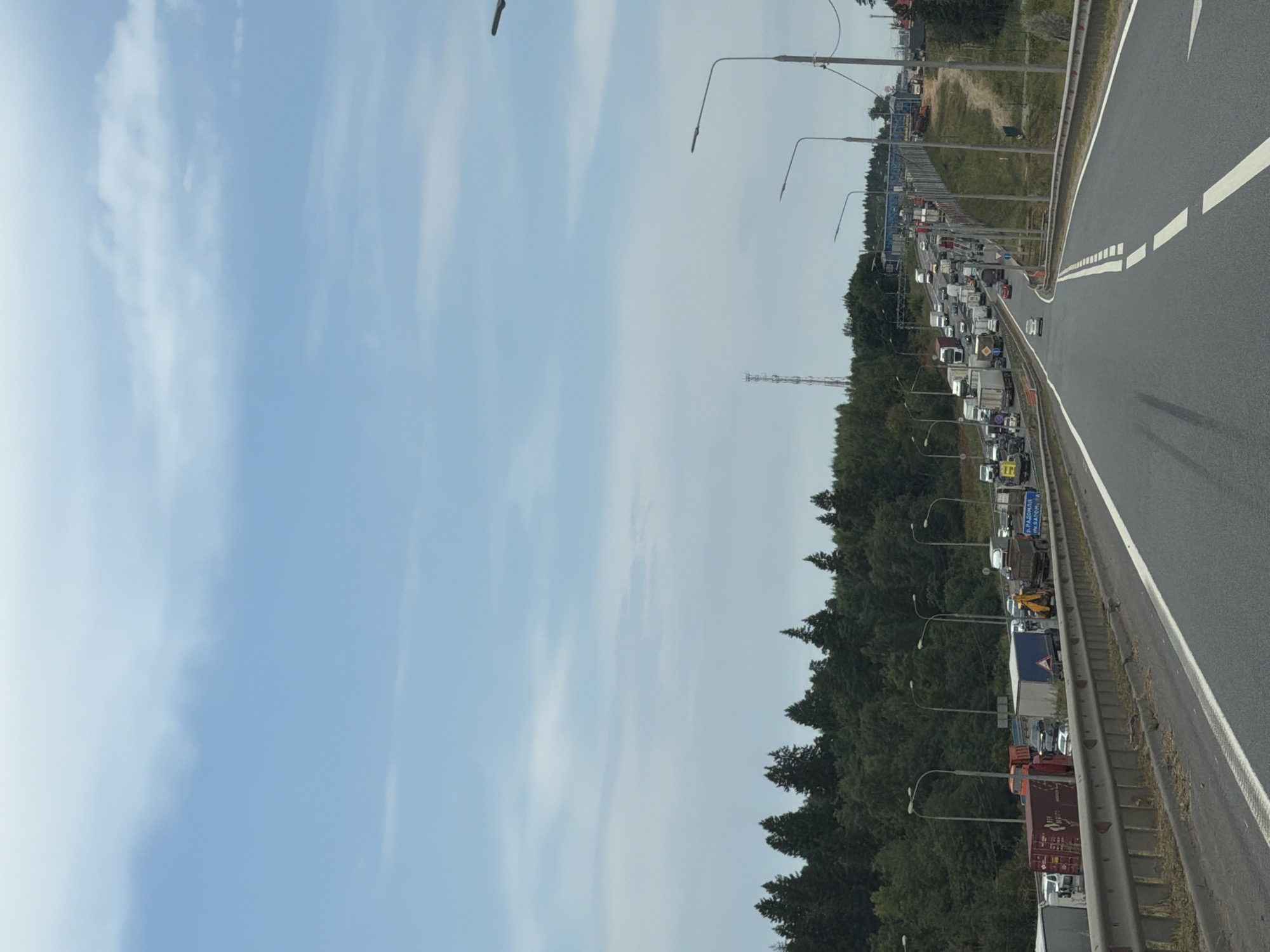 This is a photograph taken from a moving vehicle on a highway in Durykino, Russia. The highway is a multi-lane road with a guardrail on the right side. The road is congested with a significant traffic jam of trucks and cars. The vehicles are lined up for a considerable distance, stretching into the background.
On the left side of the highway, there is a dense forest with tall evergreen trees. Beyond the trees, a telecommunications tower is visible in the distance. On the right side of the highway, there are more trees and a slight incline leading to a fence and some buildings in the far background. Streetlights are positioned along the highway. The sky is partly cloudy with a light blue color. The scene suggests a typical day on a busy highway, possibly experiencing delays due to traffic or roadwork.