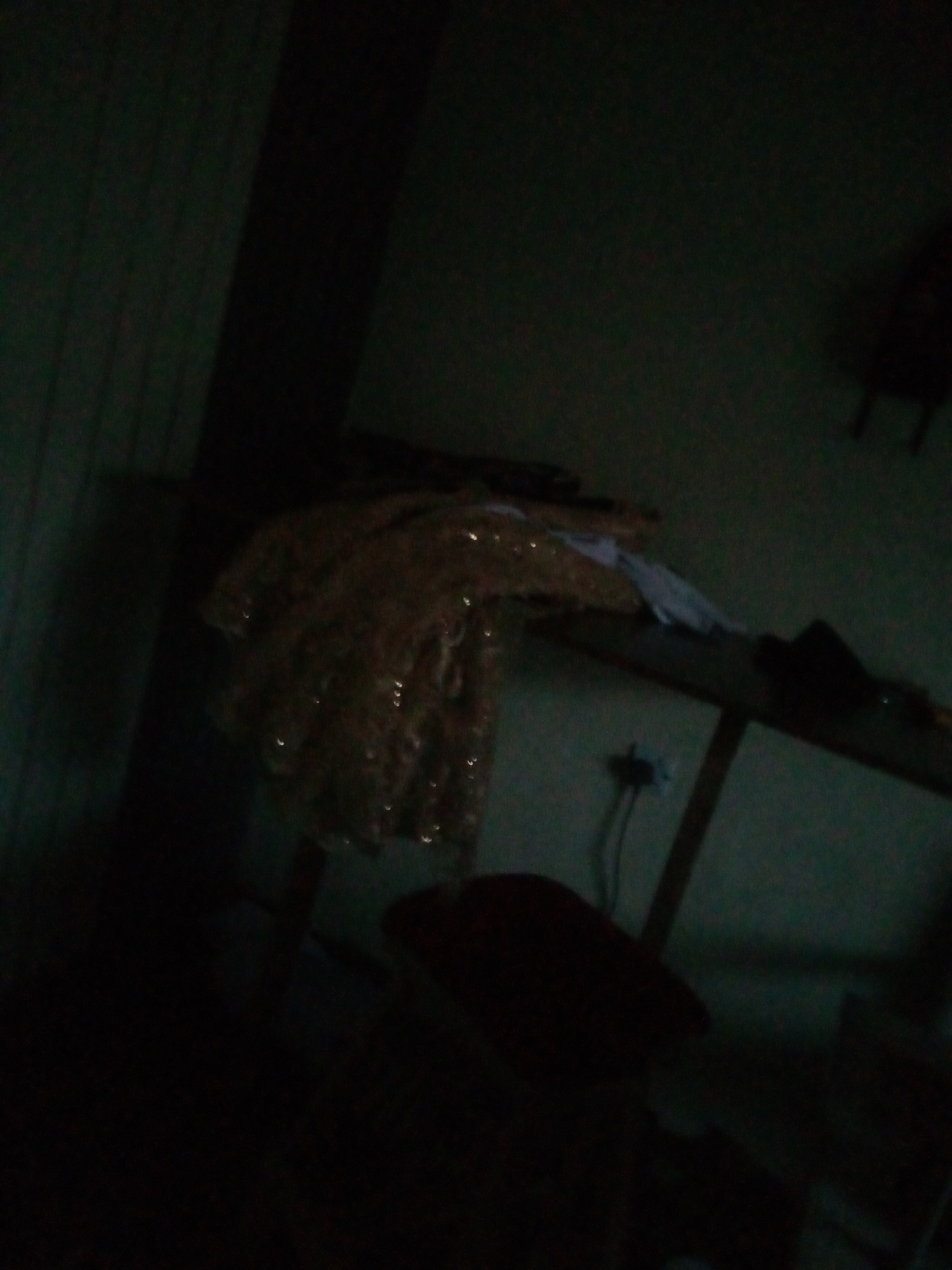 The image depicts a dimly lit interior space, likely a room in Jalingo, Nigeria. In the foreground, a dark, indistinct object, possibly a piece of furniture like a stool, is visible. Behind it, a table or shelf is present, holding various items, including what appears to be a piece of ornate fabric with reflective elements, possibly sequins or beads, draped over it. The background shows a wall with vertical lines, suggesting wallpaper or paneling, and further into the darkness, another object, possibly furniture, is dimly outlined. A wall socket with a plug is also visible on the wall. The overall atmosphere is dark and somewhat cluttered, with limited visibility due to poor lighting.