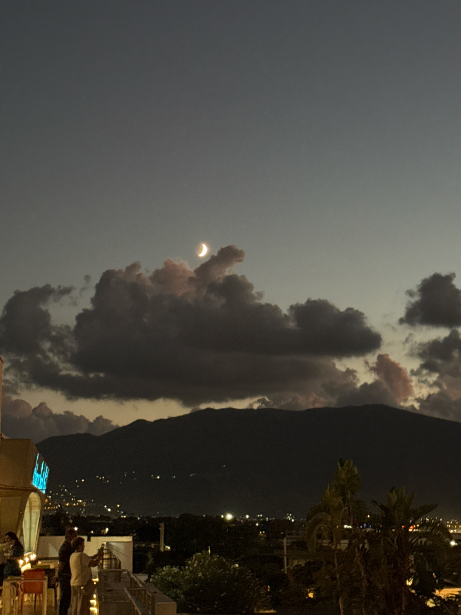 The image shows a nighttime view from a rooftop terrace overlooking the city of Isola delle Femmine, Italy. A crescent moon is visible in the dusky sky, partially obscured by dark clouds. In the foreground, several people are standing on the terrace, some appearing to be engaged in conversation. One person is holding what looks like a phone or camera. There are tables and chairs on the terrace, suggesting it is part of a restaurant or bar. The city lights twinkle in the distance, spread across the landscape and climbing up the side of a dark mountain range that dominates the background. Palm trees are also visible on the terrace. The overall atmosphere is serene and captures the ambiance of a Mediterranean evening.