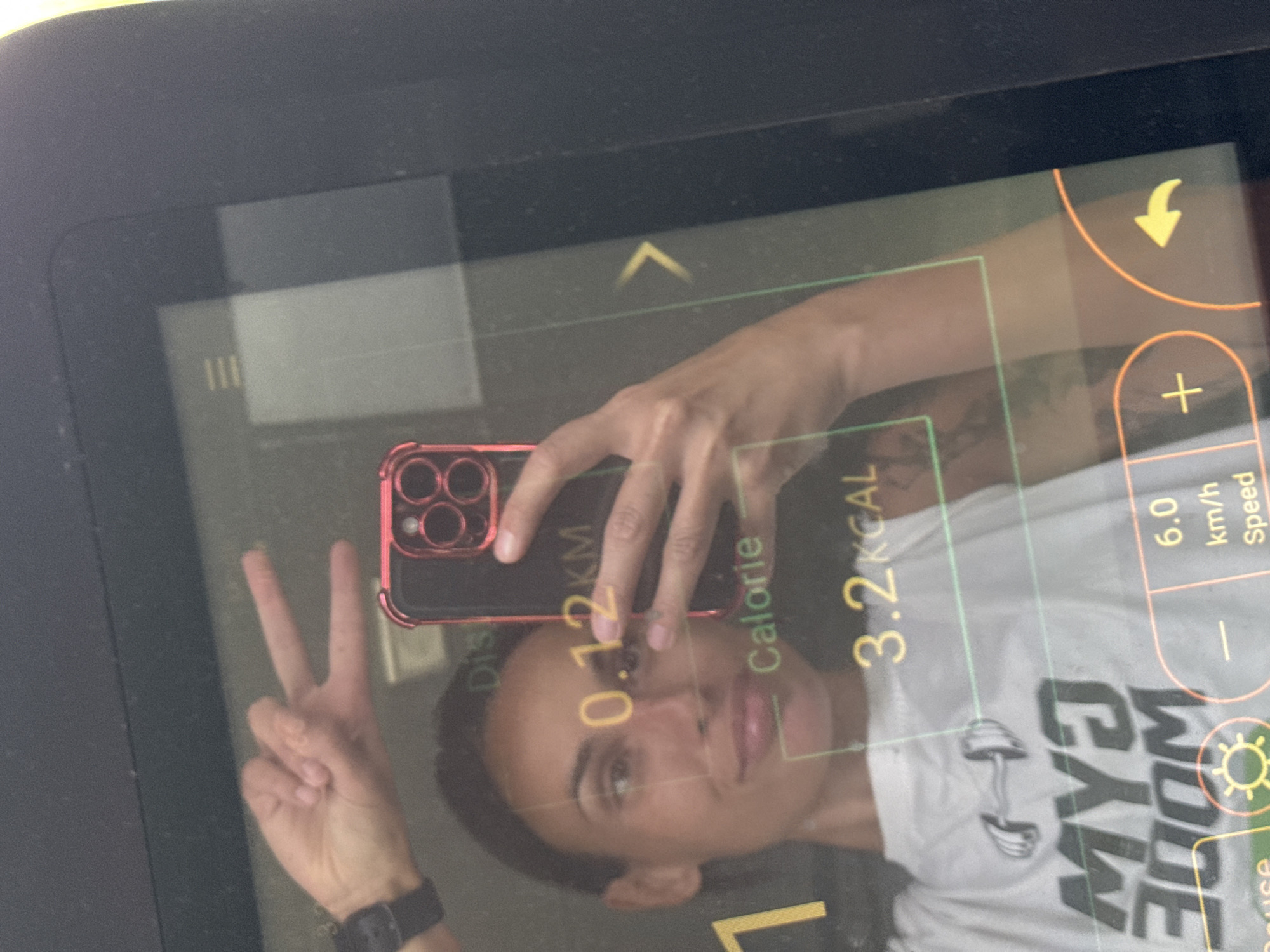 A woman is reflected in the screen of a treadmill, holding a smartphone up to take a selfie. She is wearing a white t-shirt with a graphic of a dumbbell and the text "MY3 3OOM" on it. Her left hand is raised in a peace sign, with a smartwatch on her wrist. The treadmill screen displays workout data including "0.12 KM", "Calorie", "3.2 KCAL", and "6.0 km/h Speed". The background is dark and indistinct. The image is cropped to focus on the woman and the treadmill screen.