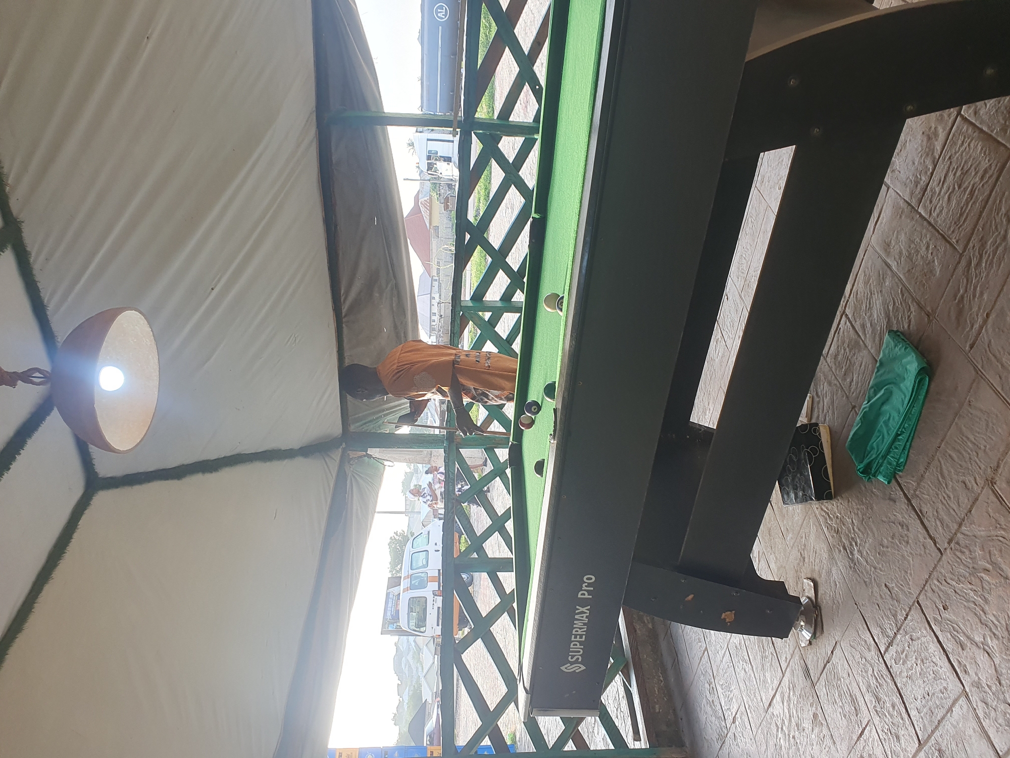 The image shows a pool table under a covered outdoor area in Lokoja, Nigeria. A man in an orange shirt is standing behind the pool table, holding a cue and appearing to play. Several billiard balls are scattered on the green felt surface of the table. The table itself is labeled "SUPERMAX Pro". In the background, through a green wooden fence, there are glimpses of vehicles, including a white van and a large truck, and buildings, suggesting an urban or semi-urban setting. The ground is tiled. A green cloth is folded on the floor near the pool table. A light bulb hangs from the ceiling of the covered area.