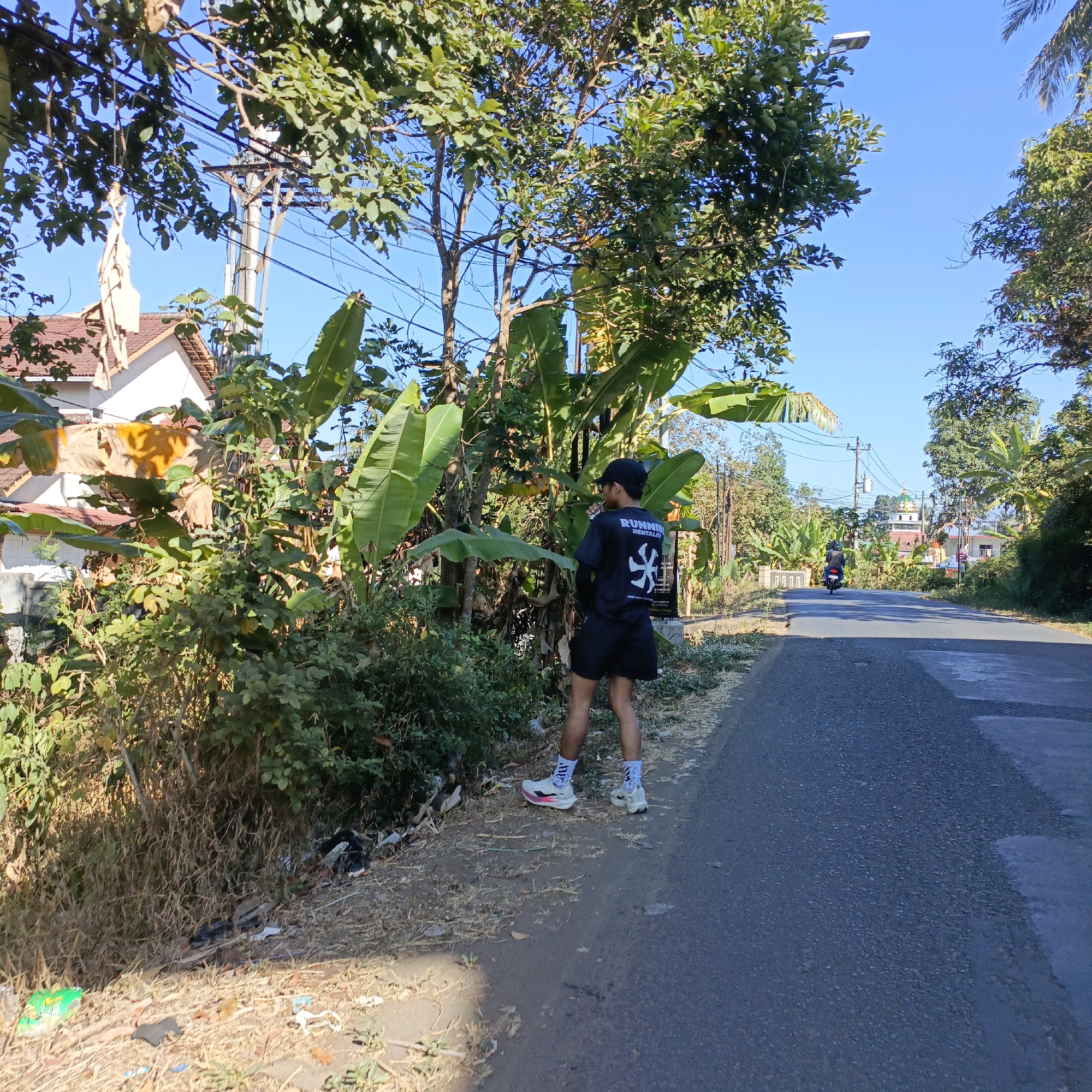 The image depicts a roadside scene in Candirejo, Indonesia, on a clear day. A young person wearing a black t-shirt with white graphics, black shorts, white socks, and white and pink sneakers stands facing away from the camera, near a dense patch of greenery and banana trees. They are wearing a black baseball cap. Next to the person, on the left side of the road, is a grassy verge with scattered litter. A paved road runs alongside the verge. In the background, a motorcycle is seen in the distance on the road, and further back, buildings and lush trees line the route, under a bright blue sky. The overall impression is of a sunny rural or semi-rural environment.