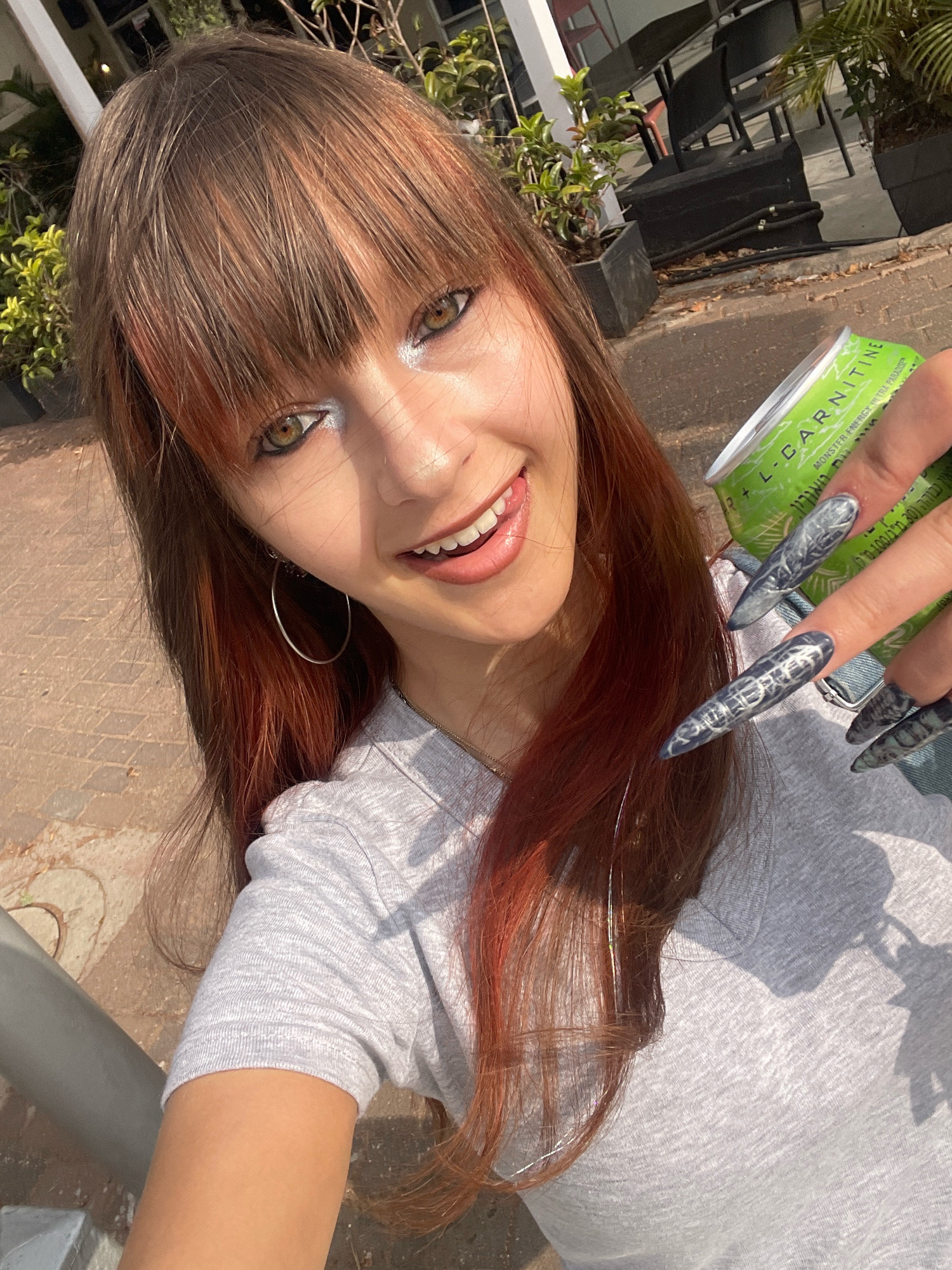 A young woman with long, reddish-brown hair and bangs is taking a selfie outdoors. She is wearing a grey t-shirt and silver hoop earrings. Her makeup includes silver eyeshadow. She is holding a green can with a label that reads "L-CARNITINE MONSTER ENERGY ULTRA PARADISE". Her fingernails are long and painted with a dark, textured design. She is sticking her tongue out slightly and smiling. The background shows potted plants and outdoor seating, suggesting a cafe or restaurant setting. The pavement in the foreground appears to be paved with square stones. The scene is likely in Raanana, Israel, given the presence of Hebrew text on the can.