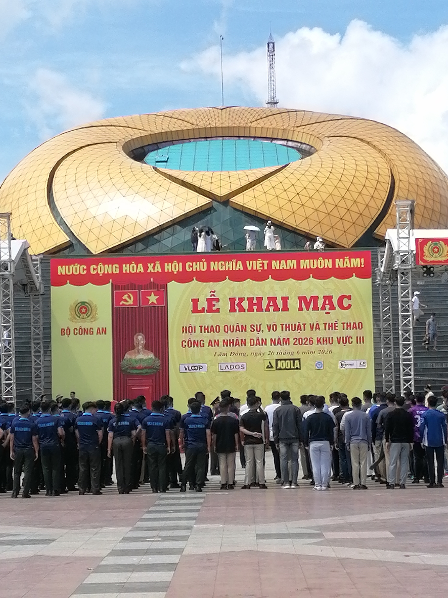 The image depicts an outdoor event in Da Lat, Vietnam, likely a ceremony or sports event. Dominating the background is a large, modern building with a distinctive golden, petal-like roof structure. In front of this building, a large banner is displayed, featuring Vietnamese text and governmental insignia, including the emblem of the Ministry of Public Security. The banner announces "LỄ KHAI MAC" (Opening Ceremony) and mentions "HỘI THAO QUÂN SỰ, VÕ THUẬT VÀ THỂ THAO CÔNG AN NHÂN DÂN NĂM 2026 KHU VỰC III" (Military, Martial Arts, and Sports Competition of the People's Public Security in 2026, Region III). Below this, further text indicates the location as "Lâm Đồng, ngày 20 tháng 6 năm 2026" (Lam Dong, June 20, 2026).
A crowd of people is gathered in the foreground, many of them wearing matching dark blue shirts. They appear to be participants or attendees at the event. Some individuals are standing in formation, while others are dispersed. The environment suggests a public square or plaza in front of the building, paved with checkered tiles. The sky is partly cloudy. The overall scene conveys a sense of an organized official event with a focus on security forces and sports.