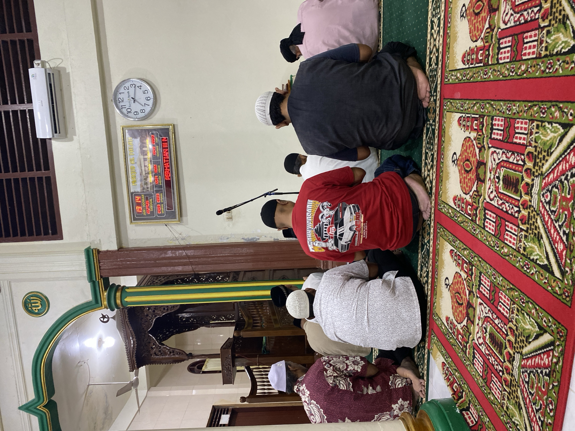 This media file captures a group of people engaged in prayer at a mosque, likely in Lhokseumawe, Indonesia. The scene is set indoors, within a prayer hall adorned with a patterned carpet featuring red, green, and beige motifs. Several individuals are visible, dressed in modest attire suitable for prayer, including skullcaps and long-sleeved shirts. Some are in the process of bowing or prostrating, indicating the ongoing prayer.
Prominently displayed on the wall is a digital clock and calendar showing the time "19 14" and the date. Below it, a framed sign reads "MASJID AL FALAH" and displays prayer times for various Islamic daily prayers like "SHUBUH," "DHUHUR," "ASHAR," "MAGHRIS," and "ISYA," along with corresponding times. A traditional analog clock is also visible, showing a time of approximately 3:30.
Other objects in the frame include an air conditioning unit mounted on the wall, a microphone stand, and decorative architectural elements of the mosque, such as a carved wooden pulpit and an archway with green and gold accents, bearing the Arabic calligraphy for "Allah." The overall atmosphere appears to be one of solemnity and devotion, suggesting a communal prayer session. The lighting suggests it is likely evening or night, given the digital clock display and the indoor setting.