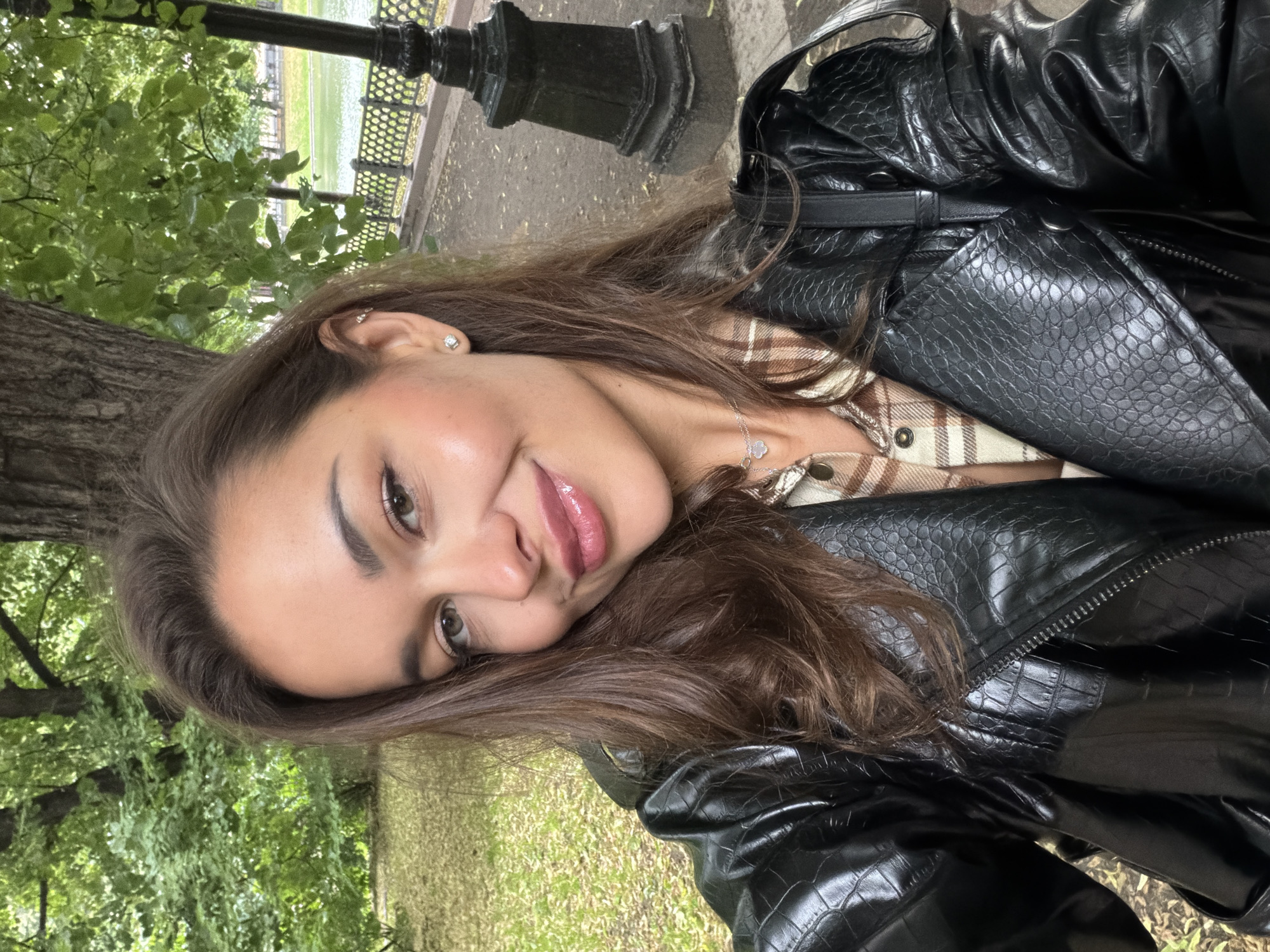 The media file is a portrait of a young woman taken outdoors, likely in a park in Moscow, Russia. She is positioned in the center of the frame, with her head tilted and her face turned towards the camera. She has long, brown hair that falls around her shoulders. She is wearing a black leather jacket with a crocodile-embossed texture over a plaid shirt. A silver necklace with a small pendant is visible on her chest.
The background suggests a natural setting with lush green trees and foliage on the left side of the image. On the right, a metal fence and a black lamppost are visible, hinting at a park or a boulevard. The ground appears to be covered with fallen leaves and grass, indicating it might be autumn or late summer.
The woman is smiling and looking directly at the camera, conveying a friendly and approachable emotion. Her makeup is subtle, with emphasis on her eyes and lips. The lighting suggests it's daytime, possibly overcast or with diffused sunlight, as there are no harsh shadows. The overall mood is casual and relaxed. There is no visible text in the image.