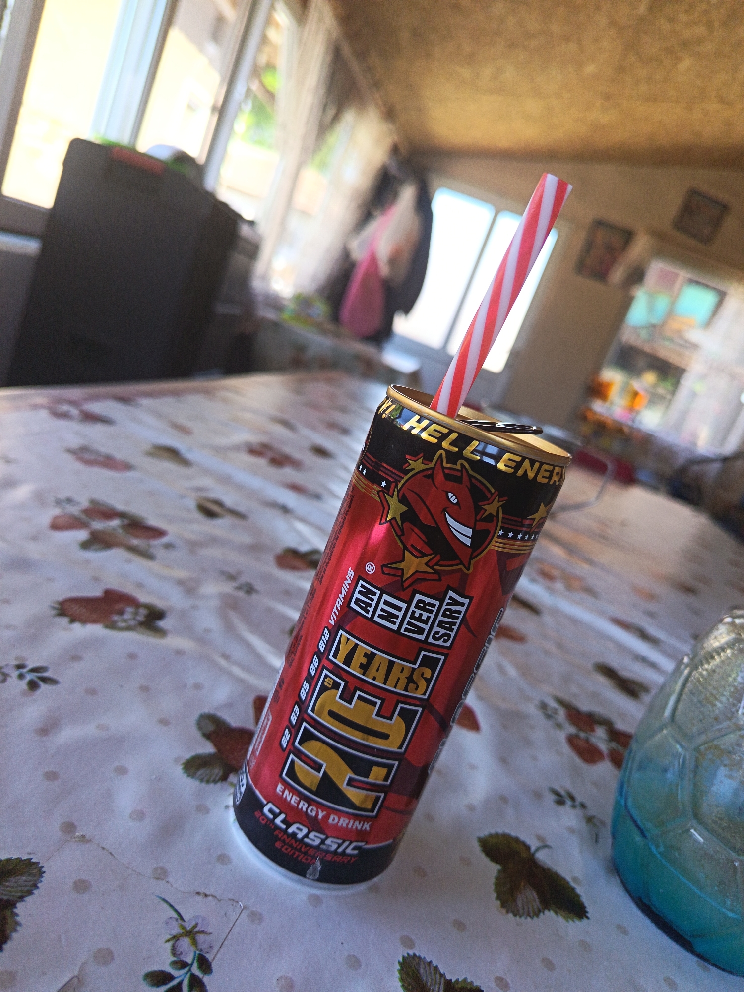 The image shows a can of "Hell Energy Classic" drink with a red and white striped straw inserted, placed on a table with a floral tablecloth. The can is red and black with gold lettering. Part of a decorative blue glass item is also visible on the table. The background is out of focus but suggests an interior space, possibly a sunroom, with windows and other furniture. The location is difficult to discern definitively, but I can note it is the city of Drenovets, Bulgaria.