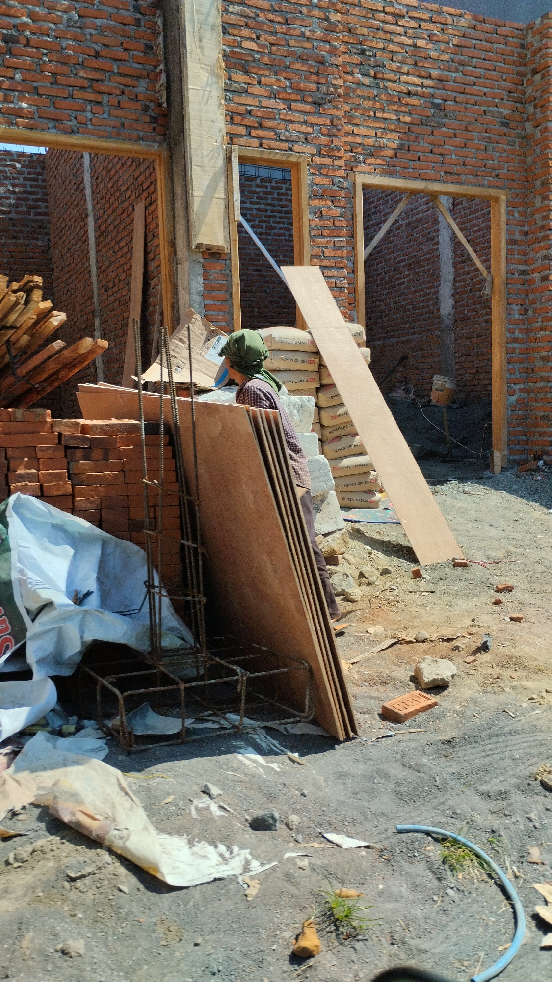 A construction site in Sidodadi, Indonesia, is depicted in this vertical, eye-level shot. A person wearing a green headscarf and a plaid shirt is seen from the side, standing near a pile of building materials. The foreground shows a heap of bricks, a large white bag, and a metal rebar cage, all resting on a dirt and gravel surface. Behind these items, stacks of plywood and bags of cement are visible, leaning against the unfinished brick wall of a building. The building features openings for doors and windows, with wooden frames in place. The ground is uneven, with scattered rocks and debris. Sunlight casts shadows, indicating an outdoor daytime scene.