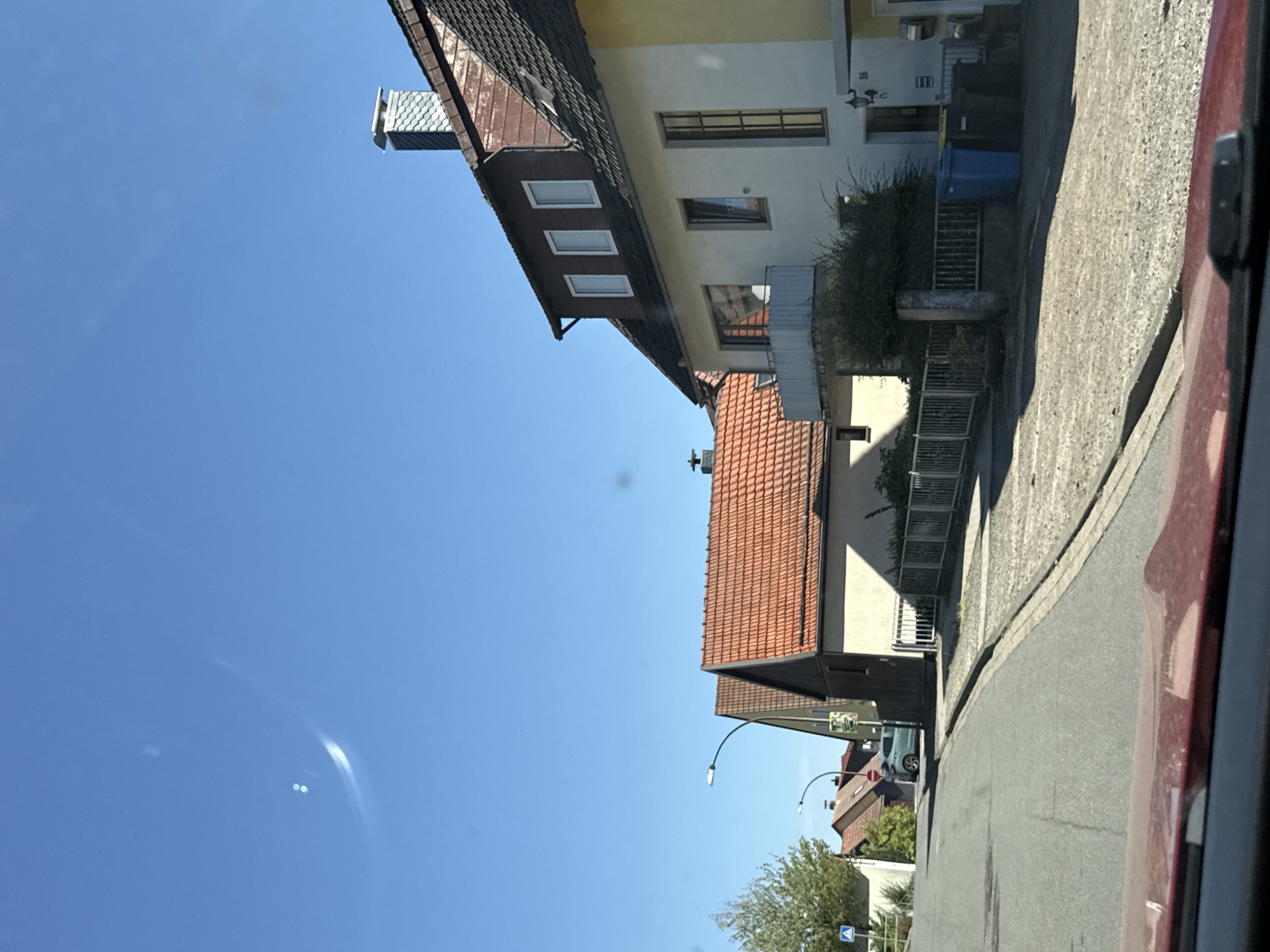 The image depicts a street scene in Bad Sachsa, Germany. In the foreground, the red hood of a car is visible. The street is paved and curves to the right. On the right side of the street, there are several houses. One house is white with a balcony and a dark tiled roof, featuring a prominent chimney. Adjacent to it, another house has a bright orange tiled roof. Further down the street, more houses with tiled roofs and trees can be seen. A light blue car is parked on the left side of the street. The sky is clear and blue. There are also several trash bins near the houses on the right.